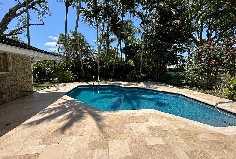 Residential backyard pool with surrounded by trees and tropical plants, stone patio, and a house wall on the left.