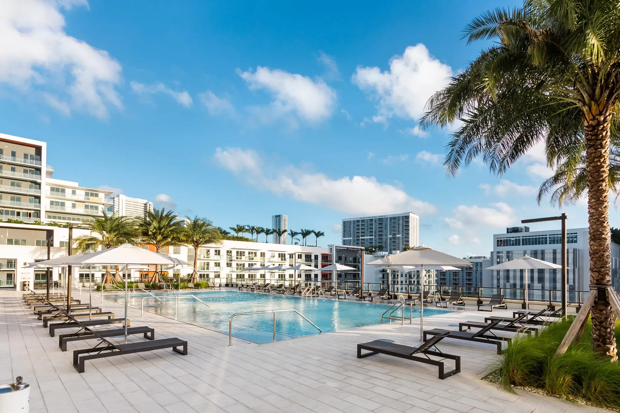 Outdoor swimming pool area with lounge chairs, umbrellas, and palm trees, surrounded by modern high-rise buildings under a partly cloudy sky.