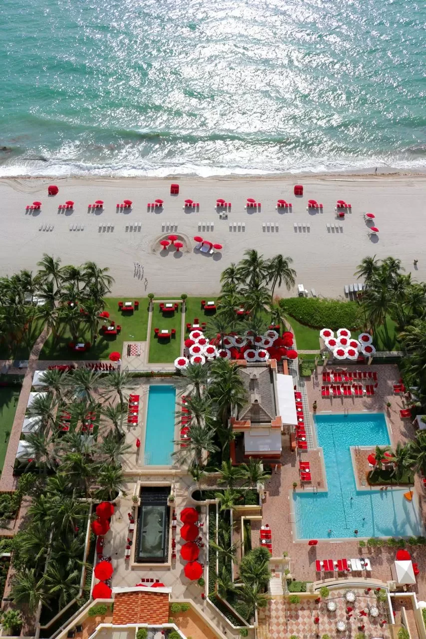 Aerial view of a beach resort with red umbrellas, lounge chairs, pools, and palm trees overlooking the ocean.