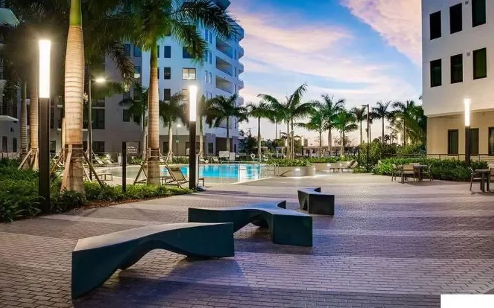 Urban outdoor pool area with palm trees, modern buildings, benches, and lighting at dusk.