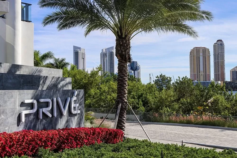 A modern cityscape with tall buildings, a large palm tree, and a sign that reads 'PRIVE' in front of a landscaped area with red flowers and green bushes.
