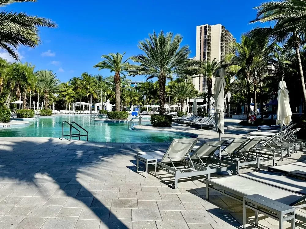 Outdoor swimming pool area with lounge chairs, umbrellas, and palm trees, in a sunny tropical location with a tall building in the background.
