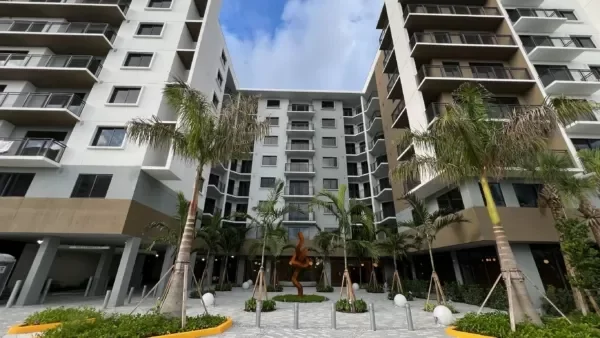 Tall white apartment buildings with balconies, palm trees, and a courtyard with a sculpture in the center.
