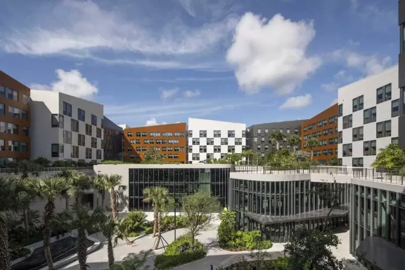 Modern residential building complex with colorful facades, landscaped courtyard with palm trees and greenery, under a partly cloudy sky.