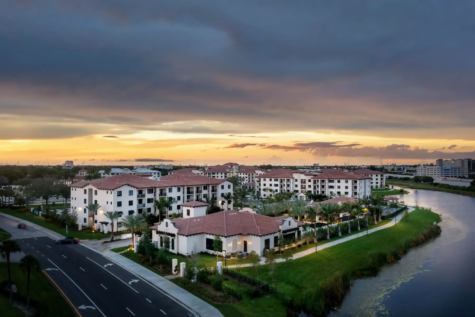 Aerial view of a residential complex with white buildings and red-tiled roofs along a waterway at sunset, with a cloudy sky above.