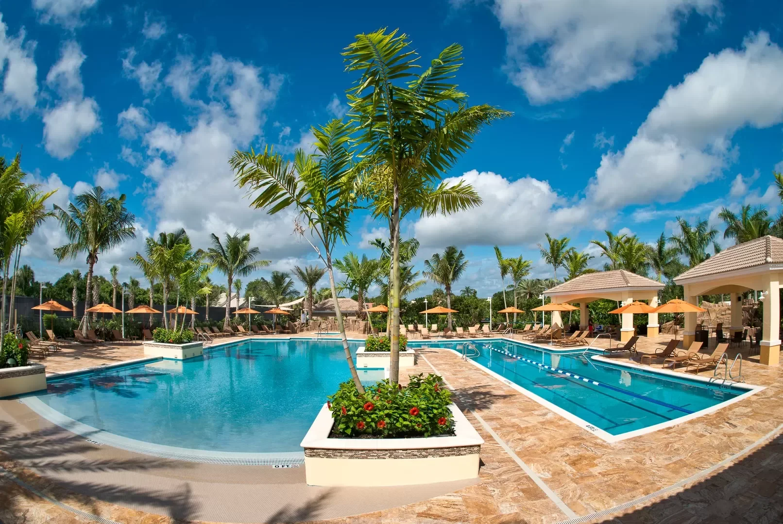 Resort swimming pool area with tall palm trees, orange umbrellas, lounge chairs, and a clear blue sky with clouds.