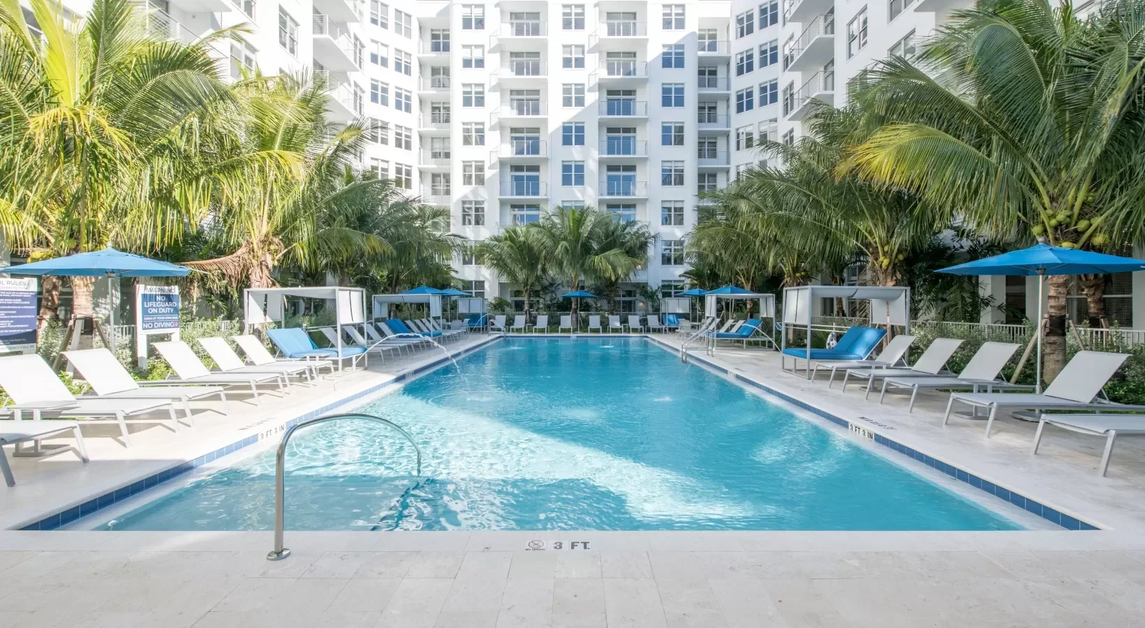 Empty swimming pool with blue lounge chairs and umbrellas, surrounded by palm trees and a tall white residential building.