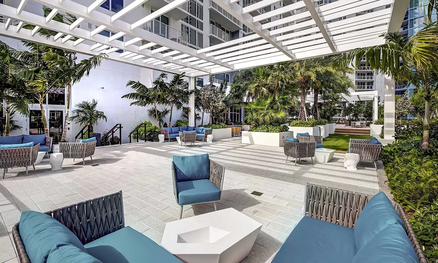 Outdoor patio area with modern furniture, blue cushions, and lush green tropical plants, partially shaded by a white pergola, in an urban setting.