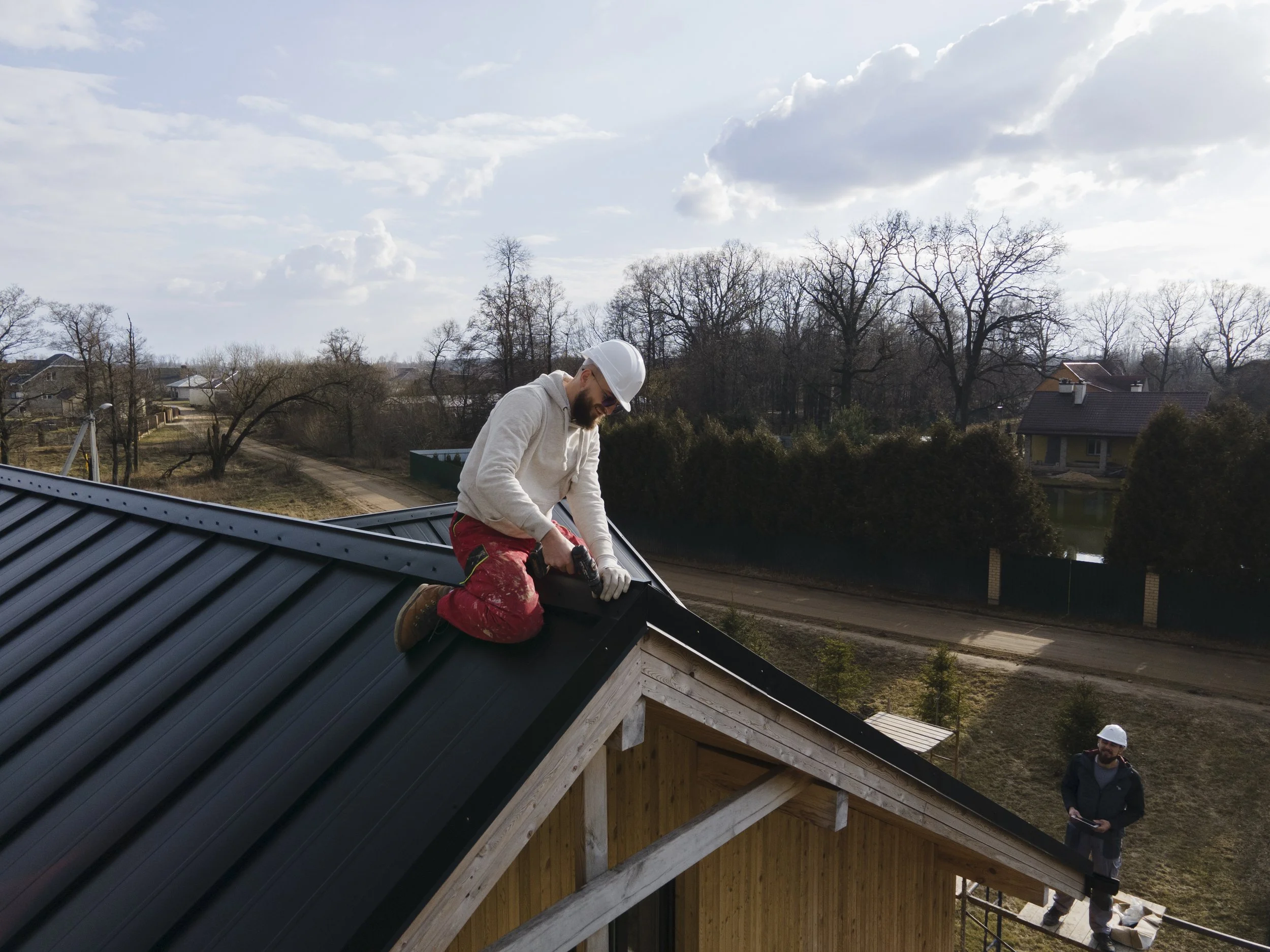 A man installing or repairing a metal roof on a house, wearing a white helmet and working with power tools, while another man with a white helmet stands below.