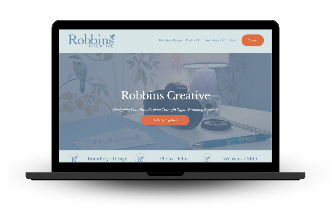 Laptop displaying a website for Robbins Creative, a digital branding service, with a blurred background of a desk, notebook, pen, lamp, camera, and wallpaper with bird illustrations.