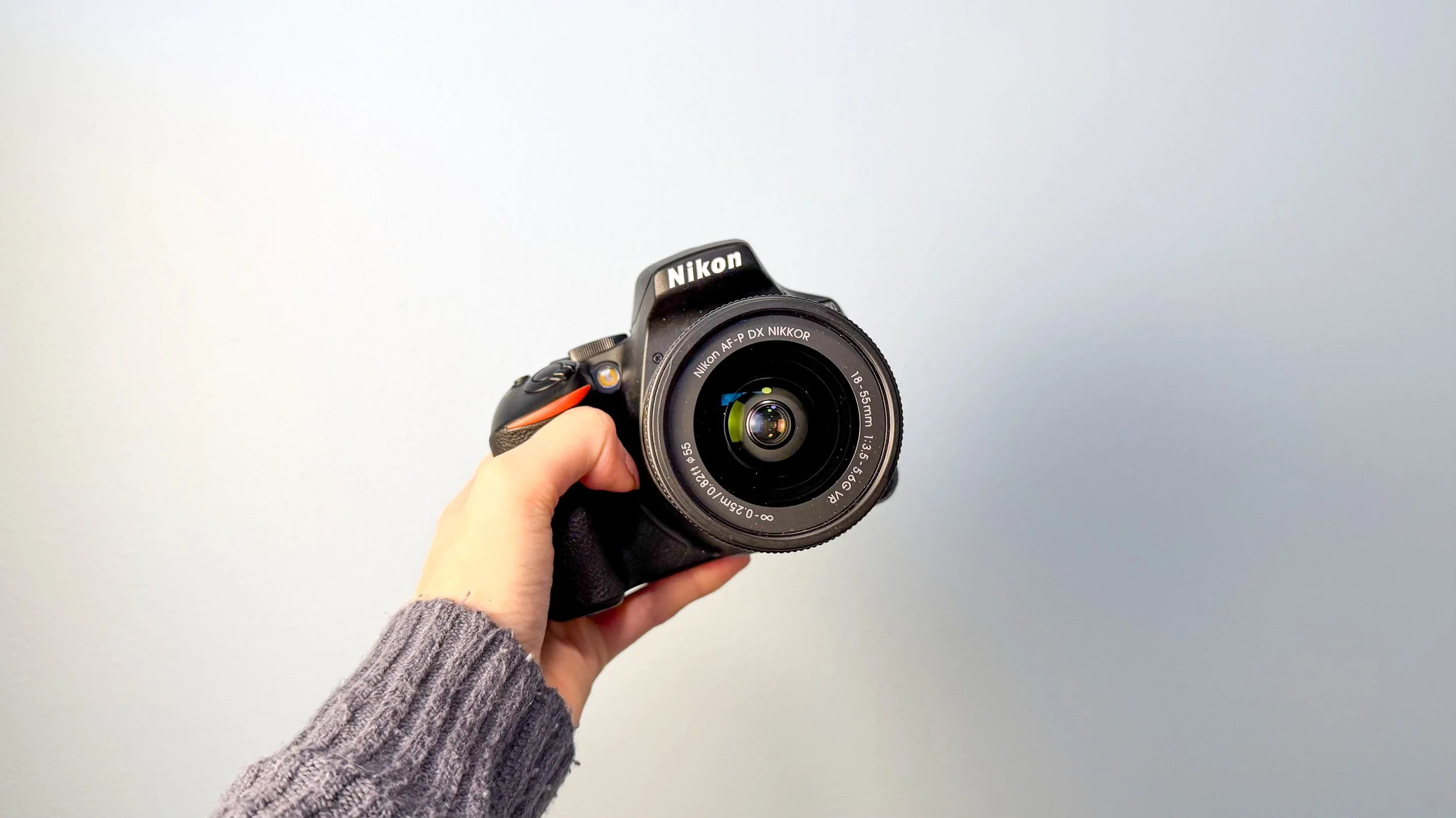 Hand holding a Nikon camera with an 18-55mm lens against a light gray background.