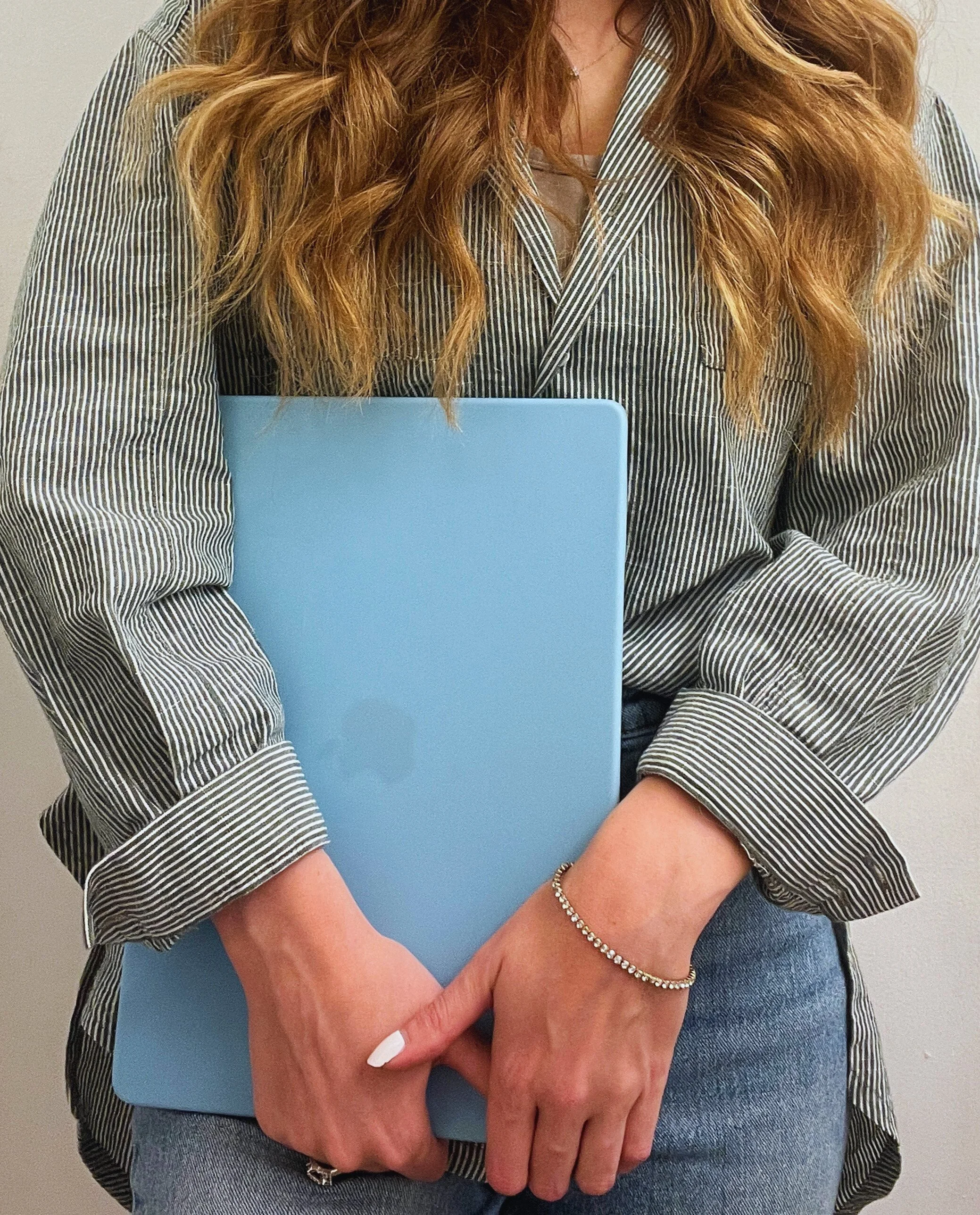 Person with long, wavy hair holding a light blue laptop, wearing a striped shirt and a bracelet.