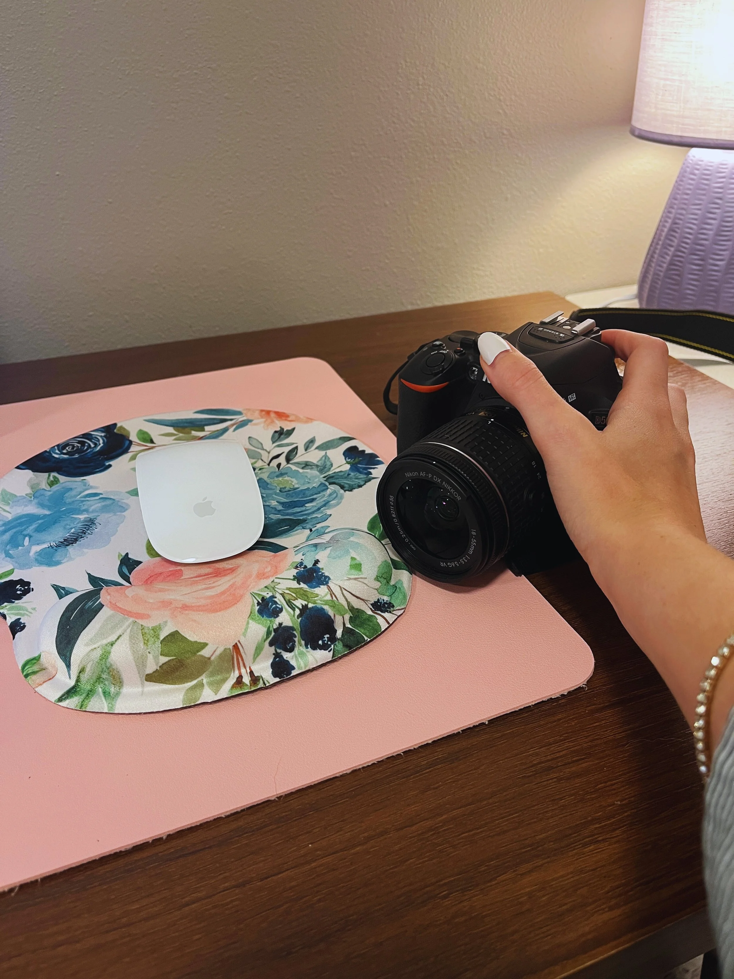 A person holding a camera next to a floral mouse pad with an Apple Magic Mouse on it, placed on a pink desk mat on a wooden surface. A purple table lamp is in the background.