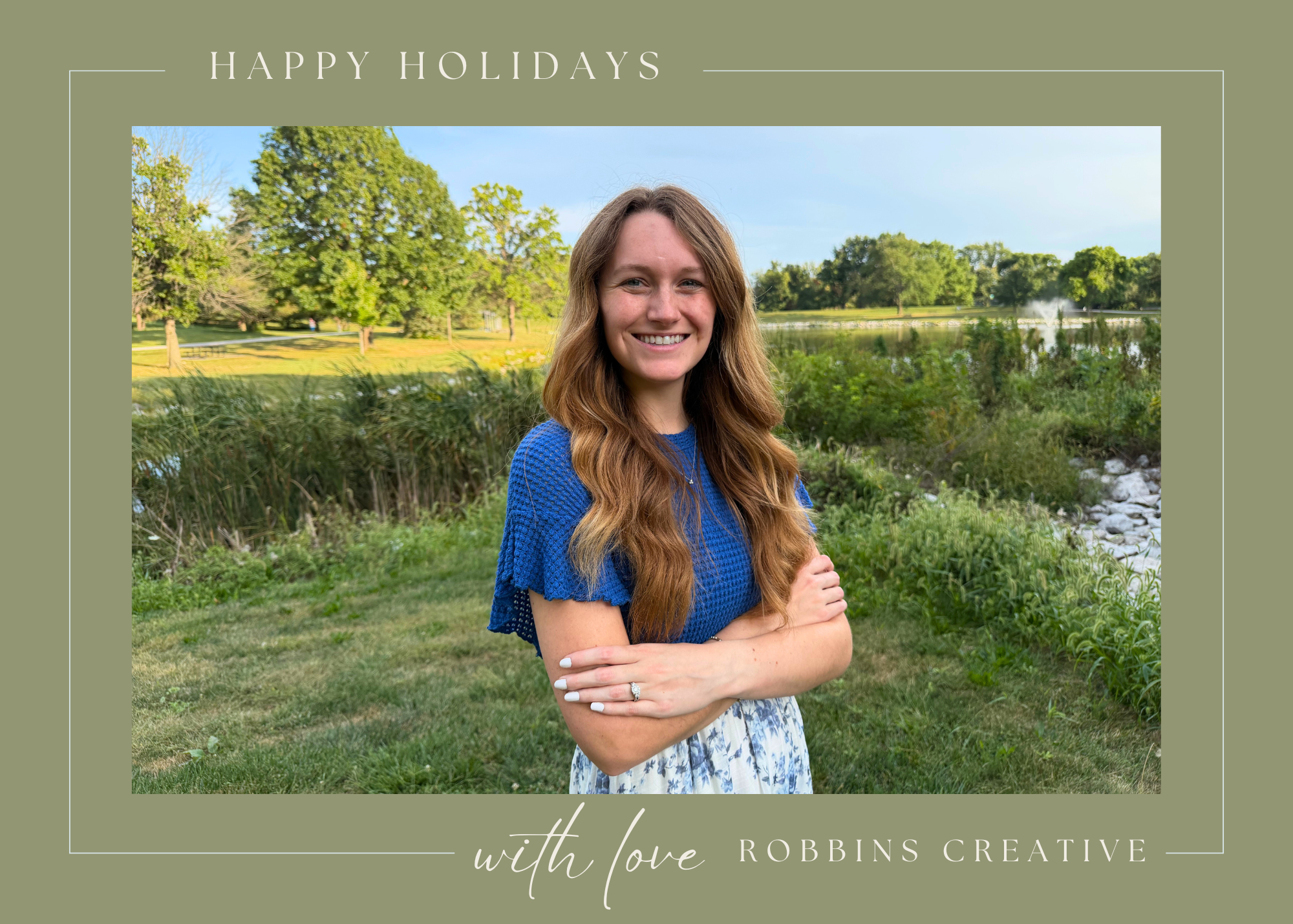 A smiling woman with long wavy hair in a blue top and floral skirt, standing with arms crossed outdoors near a lake with trees and grass, under a clear sky.