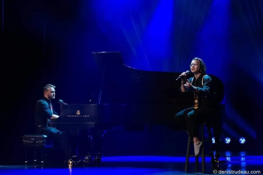 Un homme joue du piano sur scène tandis qu'une femme chante avec un micro, éclairés par des lumières bleues.