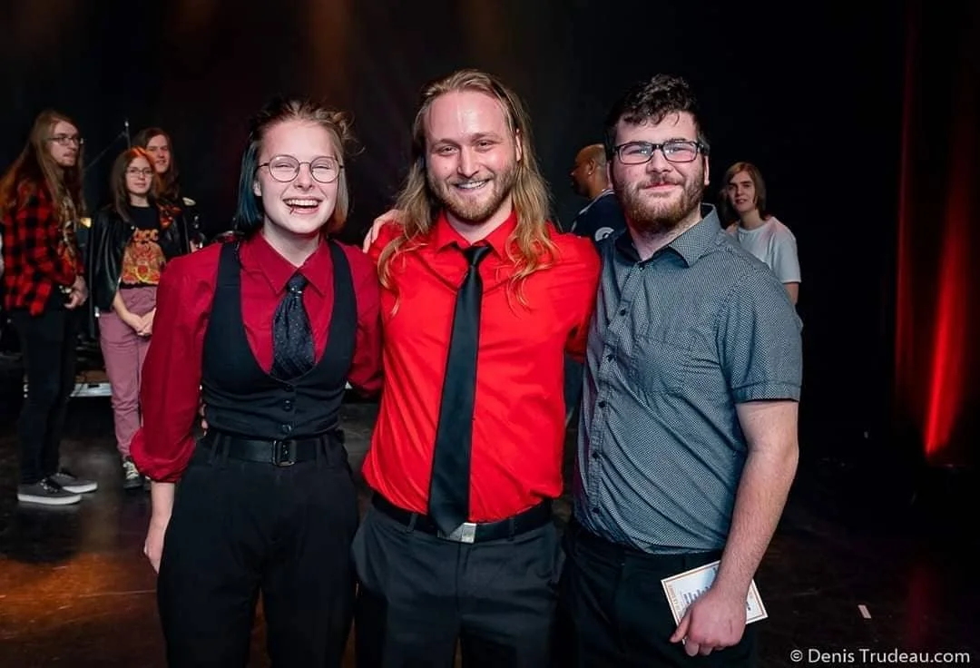 Trois personnes souriantes posent ensemble devant un fond sombre, une femme et deux hommes, à un événement.