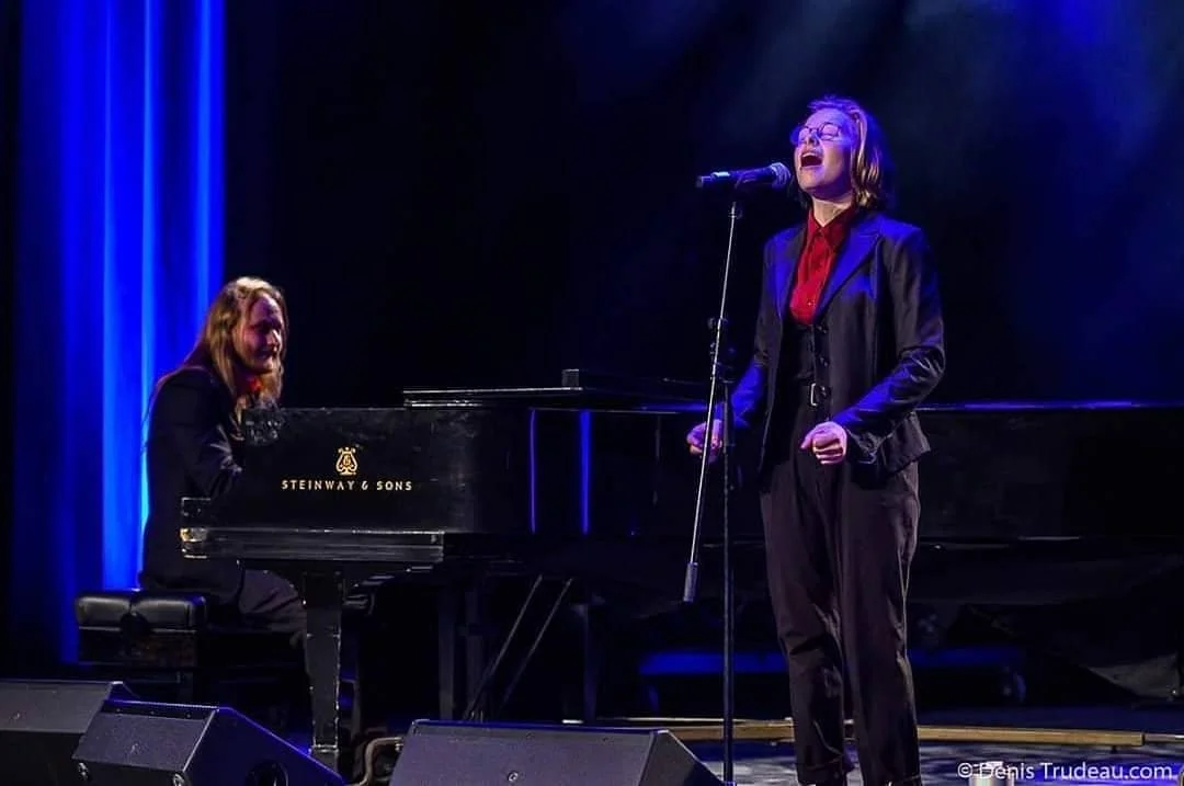 Une femme chante en tenant un micro, accompagnée d'une autre femme jouant du piano sur scène éclairée par des lumières bleues.