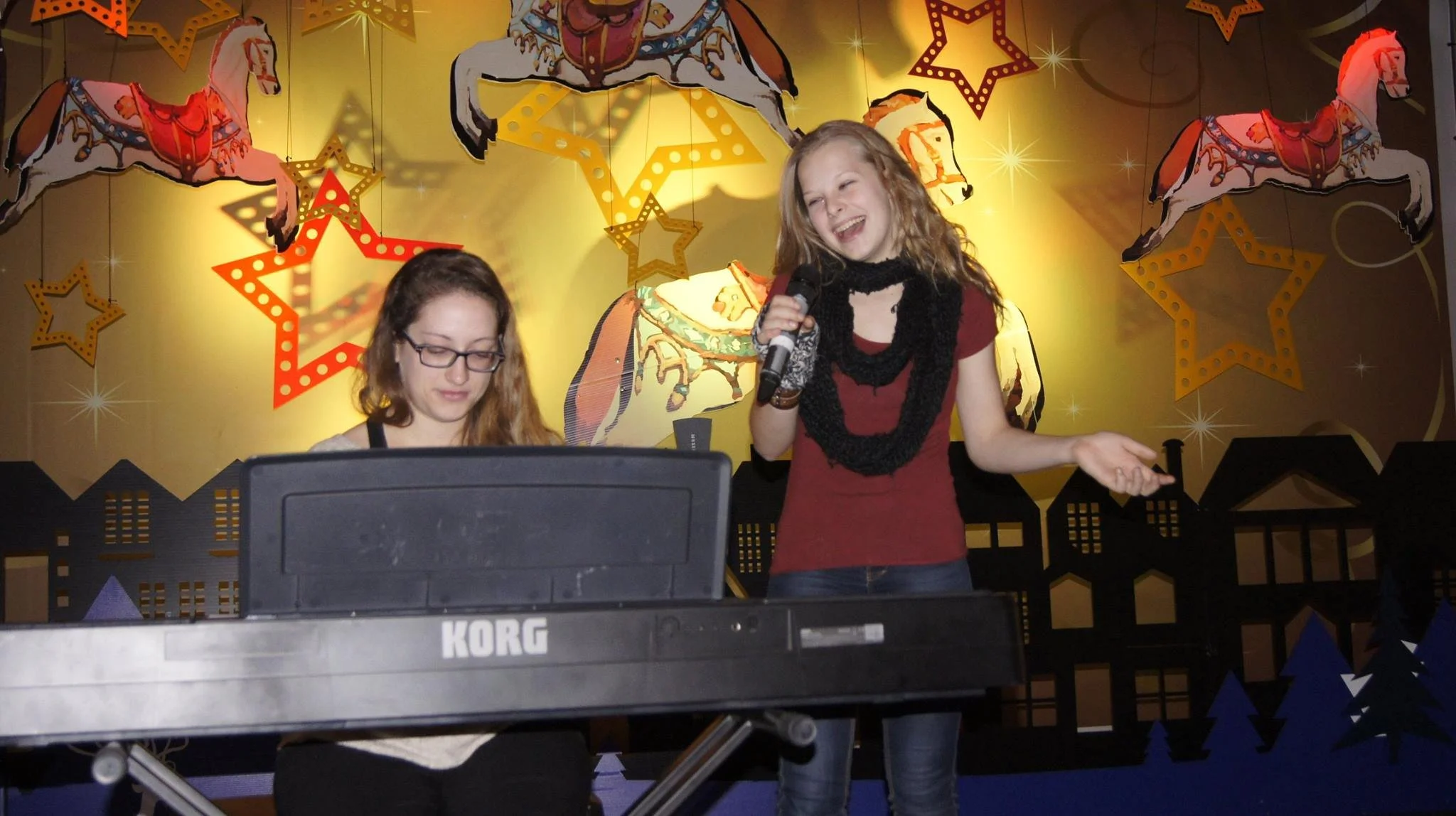 Deux jeunes femmes sur scène, l'une jouant du clavier Korg et l'autre tenant un micro, avec un fond décoré de chevaux de manège et d'étoiles lumineuses, lors d'une performance ou d'un spectacle.