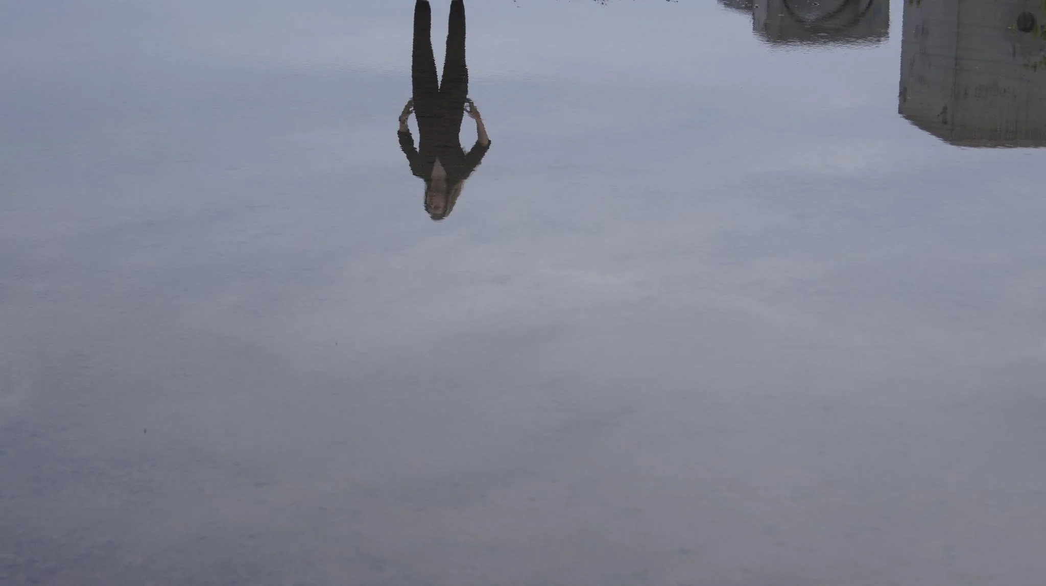 Une personne reflétée dans l'eau à cause d'une flaque ou d'une surface très calme, portant une veste noire et des lunettes, avec un bâtiment en arrière-plan.