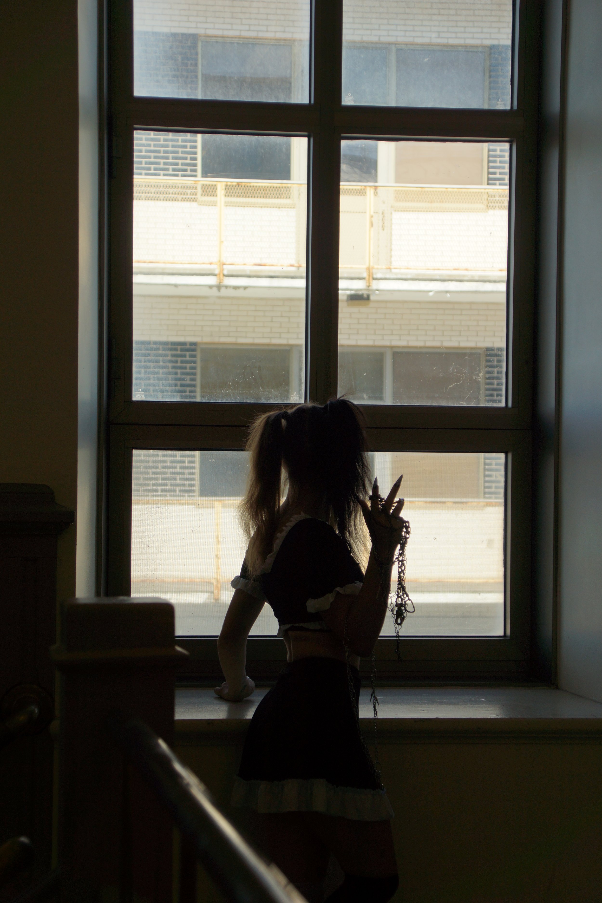 Silhouette d'une femme avec des cheveux en queue de cheval, portant une tenue noire avec des détails blancs, debout près d'une fenêtre lumineuse, tenant des bijoux ou une chaîne dans sa main.