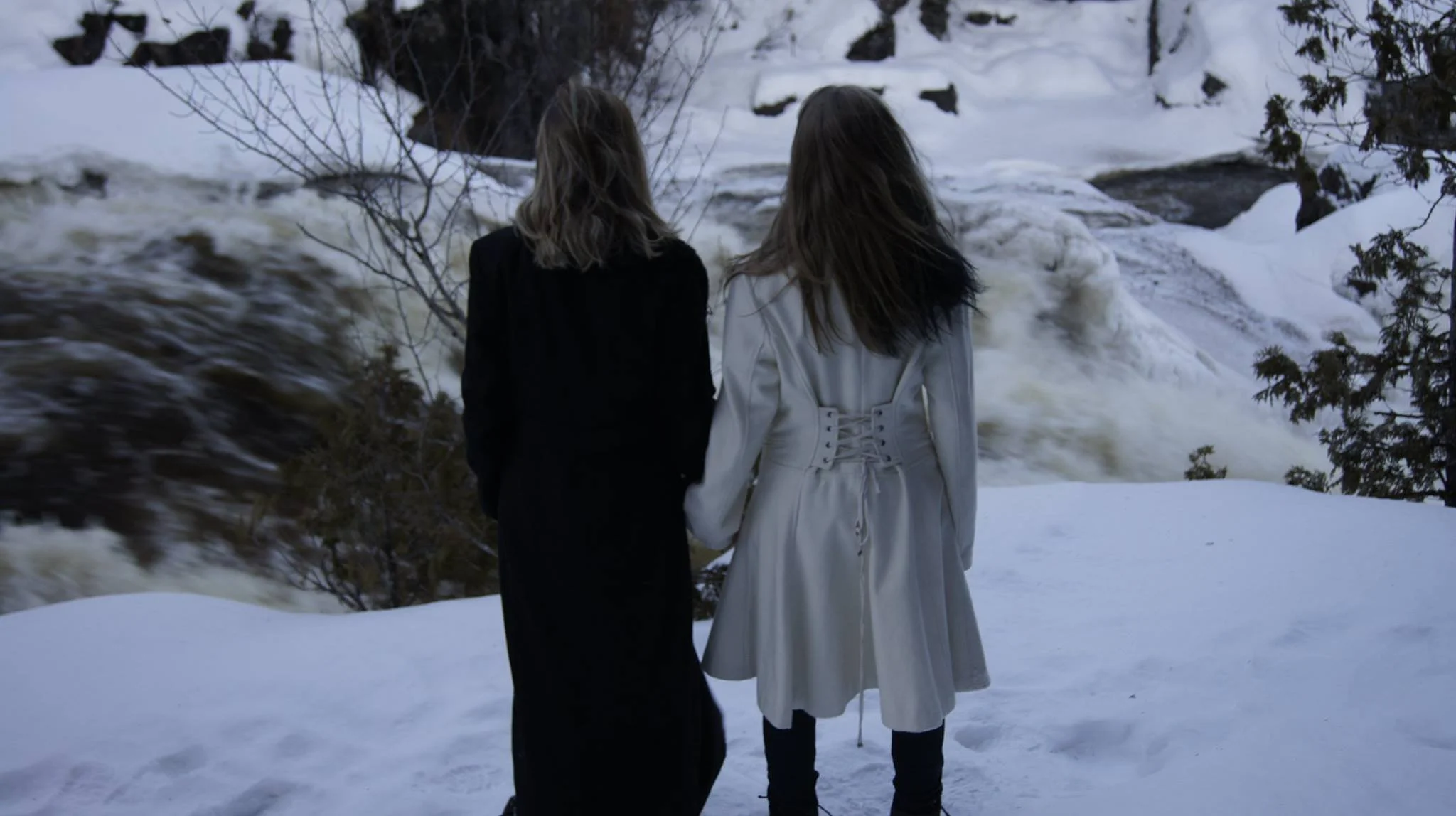 Deux femmes debout dans la neige côté à côte, regardant une cascade gelée dans un environnement enneigé.