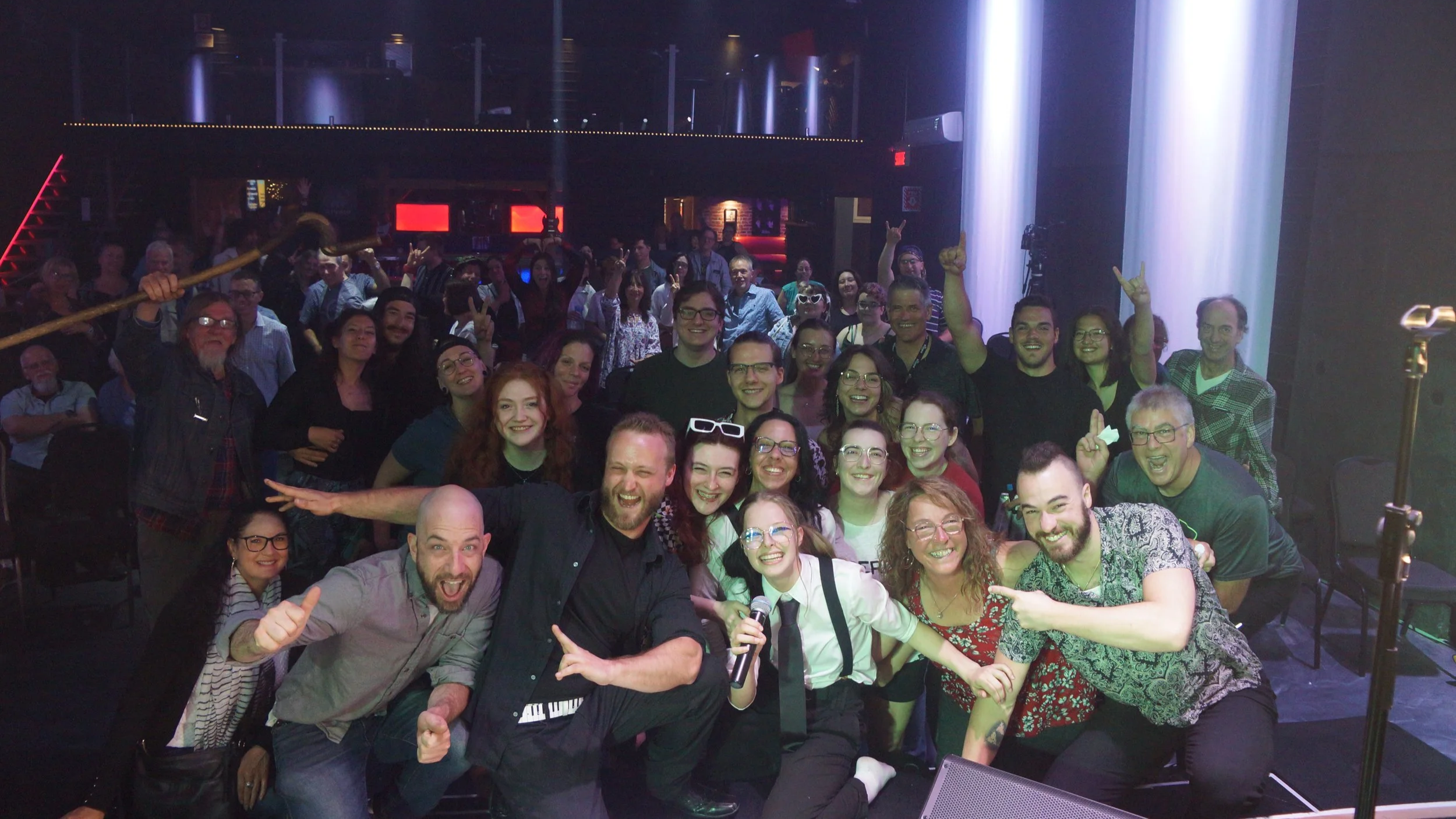 Un groupe de personnes souriantes et joyeuses lors d'une soirée ou d'un événement, posant pour une photo de groupe dans une salle éclairée avec une scène et des lumières.