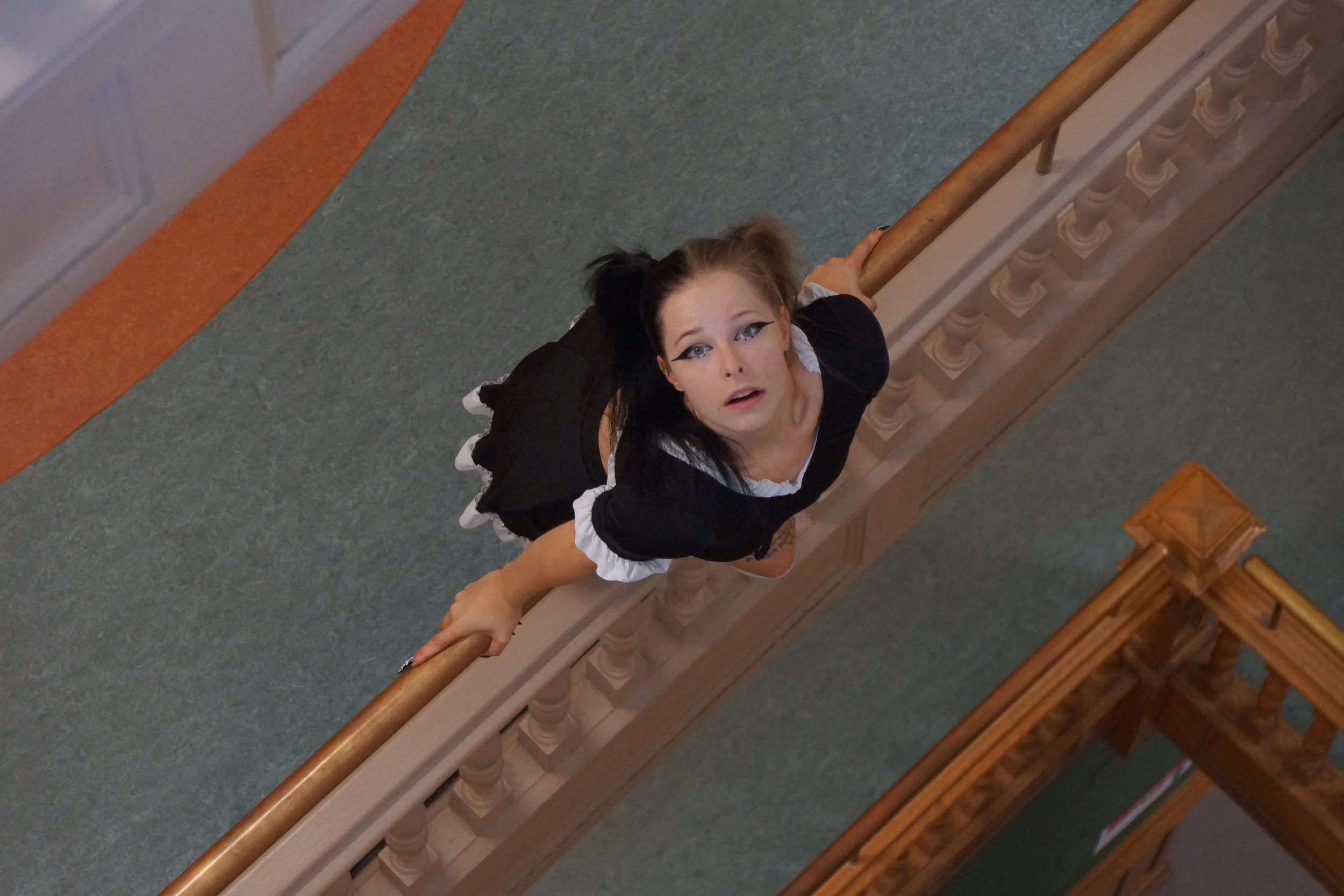 Une femme habillée en maid, vue de haut, regardant vers le haut, tenant la rampe d'escalier en bois.