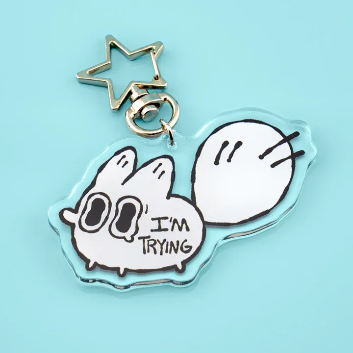 A keychain featuring a cartoon white character with large eyes and a speech bubble saying "I'm trying," attached to a gold star-shaped charm.