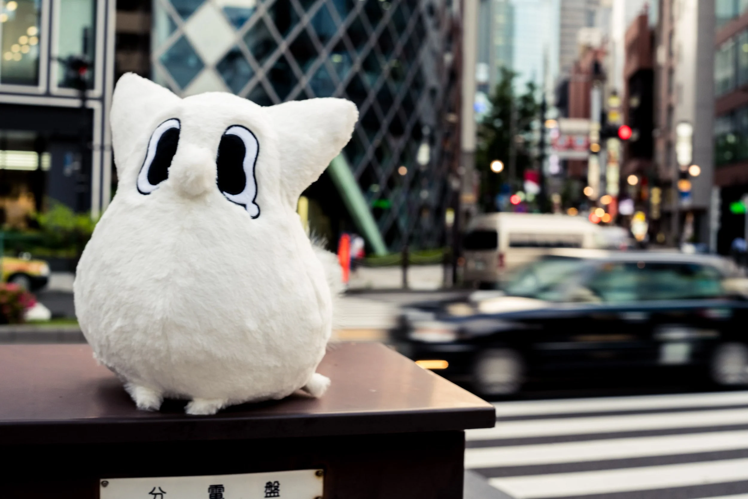 A plush toy of a white Anxiety Fox with big black eyes and tears, sitting on a wooden surface in an urban street scene with cars, buildings, and city lights in the background.