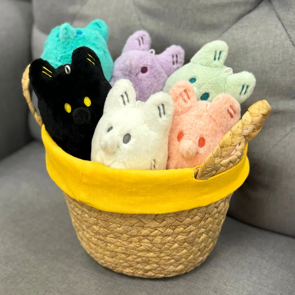 A woven basket with a yellow cloth lining filled with colorful plush animal toys from Anxiety Fox in black, white, pink, purple, and green, placed on a gray sofa.