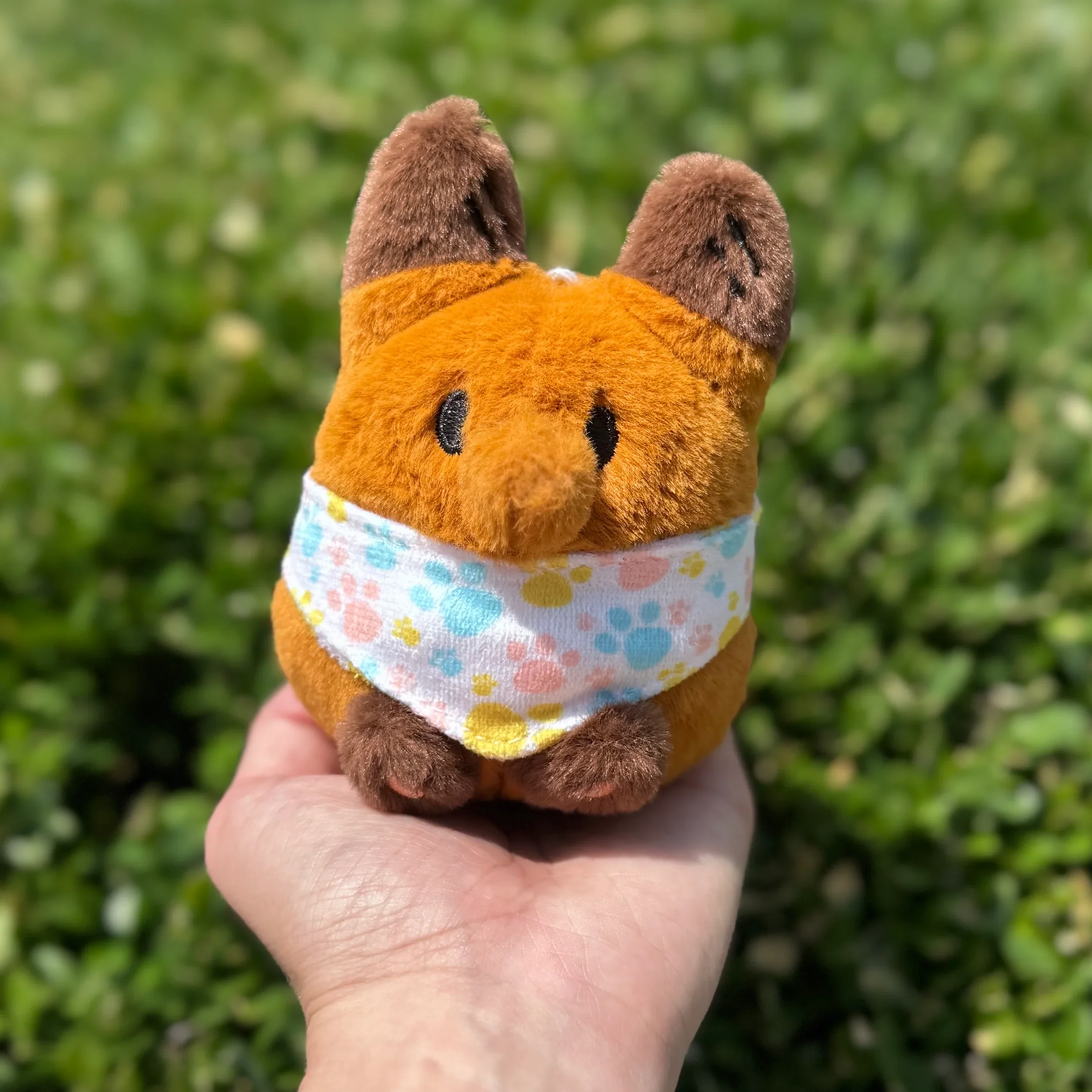 A small stuffed Anxiety Fox with brown ears, orange body, and a white bandana with colorful paw prints, held in a person's hand outdoors with green grass in the background.