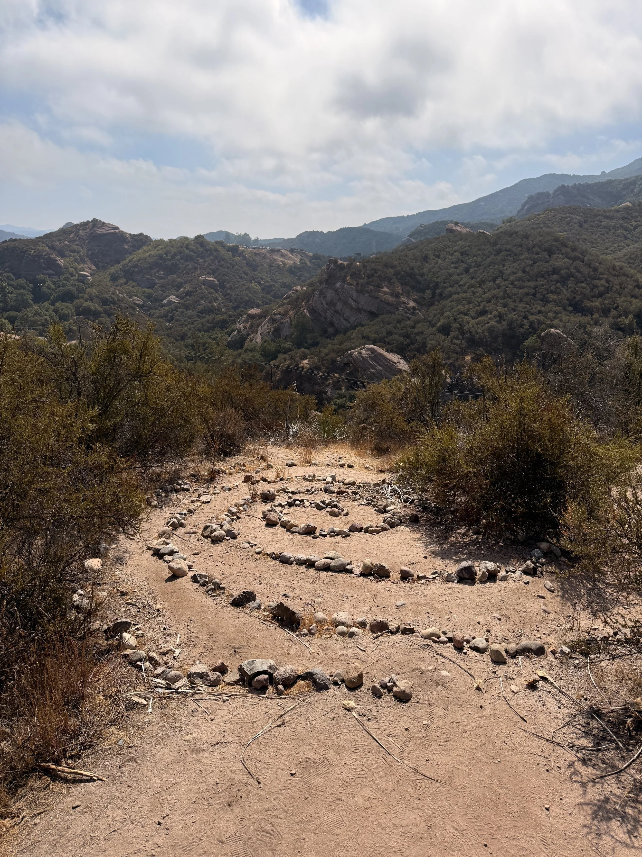 A dirt trail in Red Rock Topanga, a labyrinth made of small rocks, surrounded by desert shrubs and mountains in the background under a partly cloudy sky.