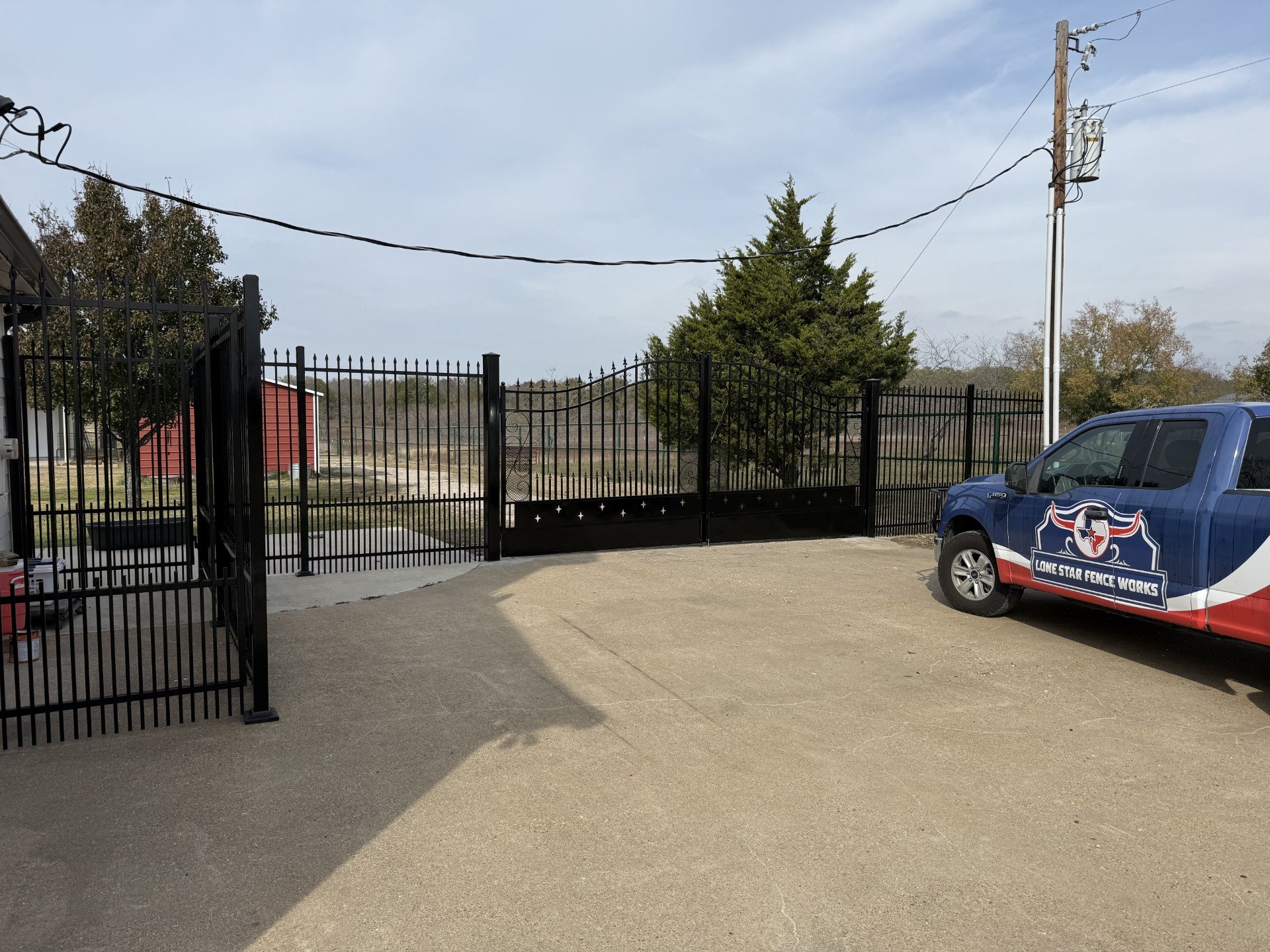 A black metal fence with a new gate encloses a concrete and dirt yard. A blue pickup truck with a logo that reads "Lone Star Fence Works" is parked nearby. A utility pole with power lines is in the background next to a large tree, with another red building and trees in the distance under a partly cloudy sky.