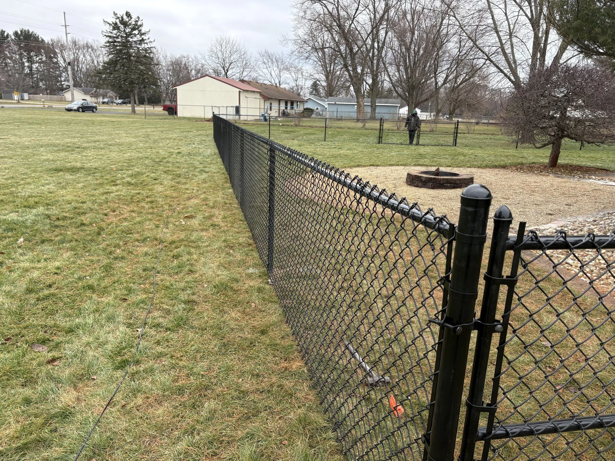 A backyard with a black chain-link fence, green grass, and a gravel fire pit. There is a person wearing dark clothing and a green hat near the fence. In the background, there are several trees without leaves, houses, and parked cars under an overcast sky.