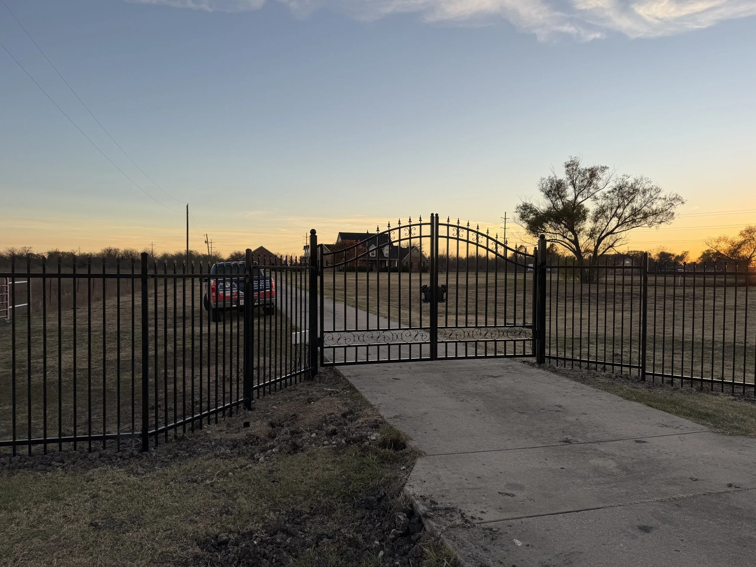 A black metal gate with decorative scrollwork leading to a large open yard at sunset, with a pathway, a tree, and a red truck parked on the left side.