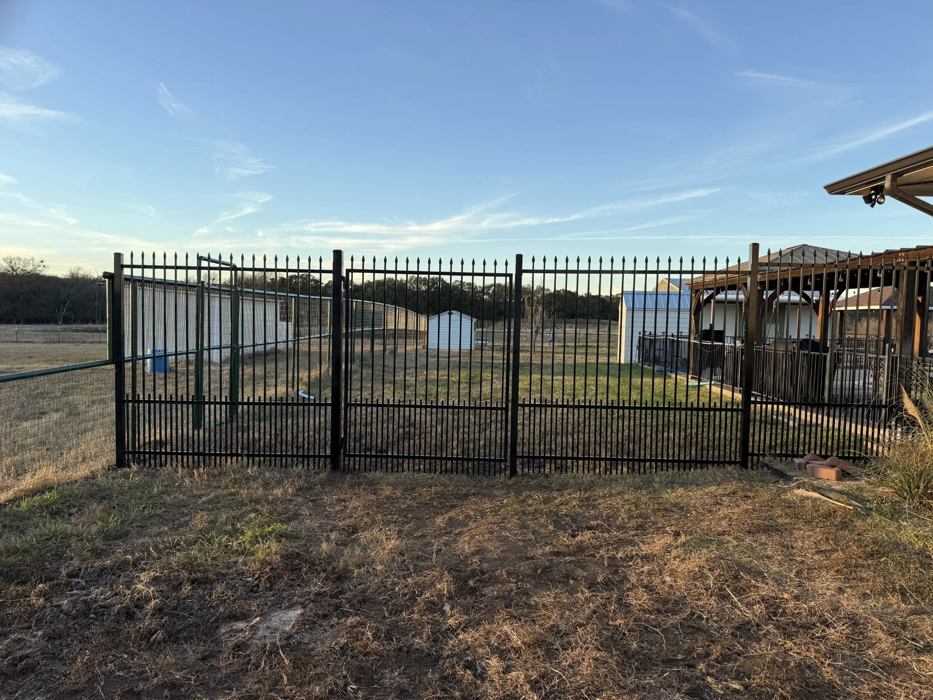 A fenced backyard with a view of a shed, greenhouse, and patio with a covered seating area under a clear sky.