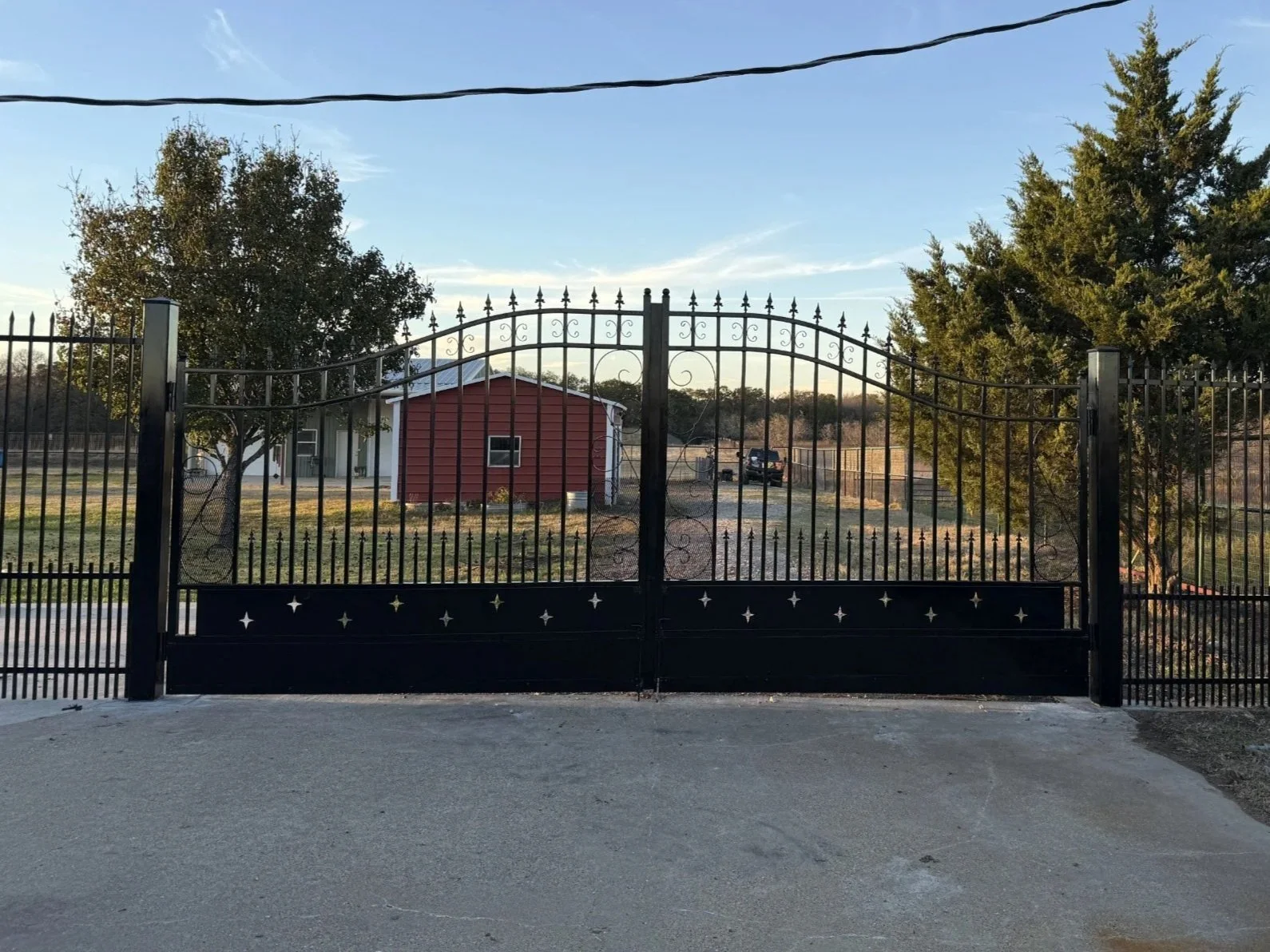 A black metal gate with decorative details in front of a rural property with trees and a red house in the background, with a small dirt parking area and a vehicle.