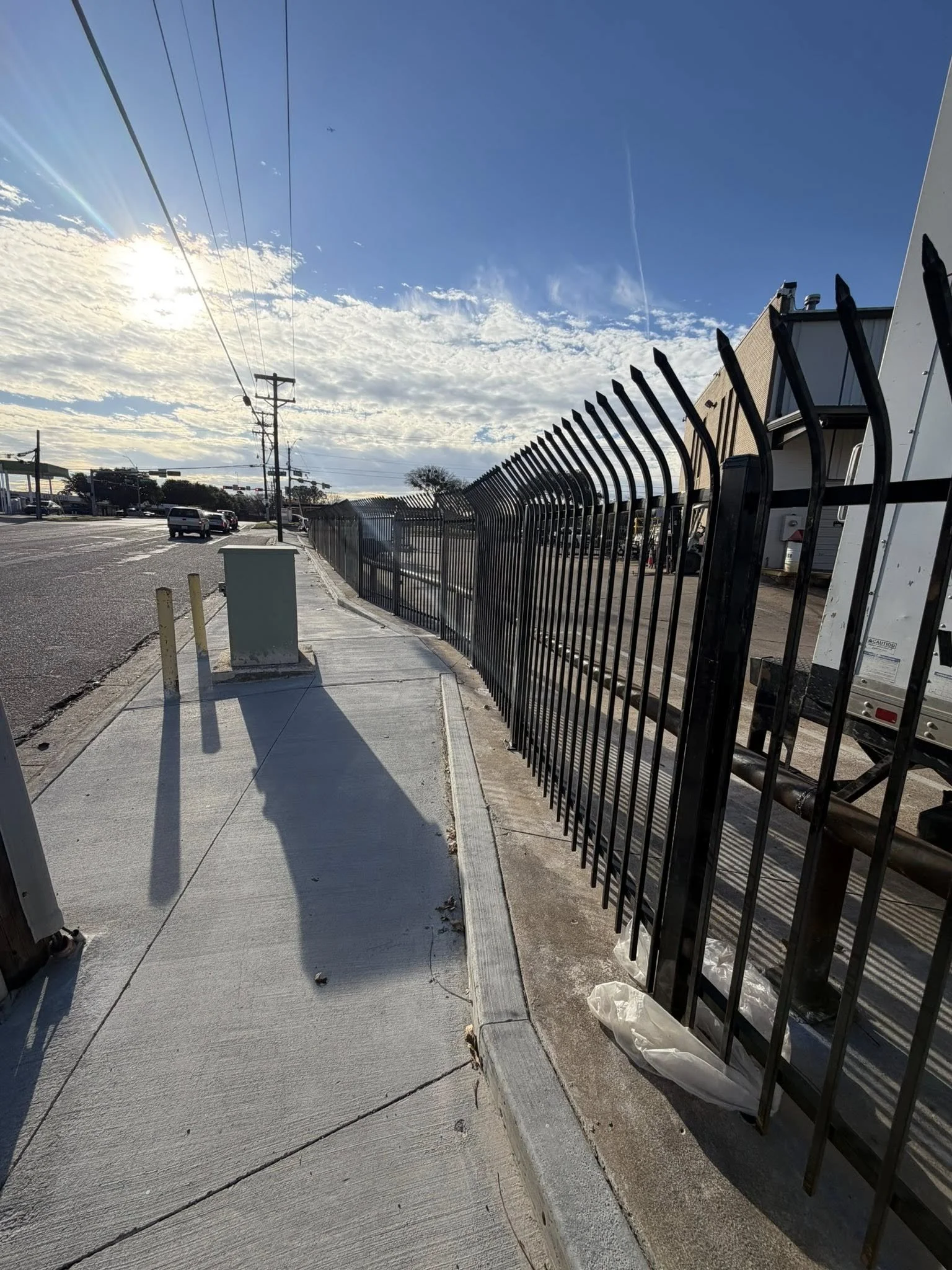 A sidewalk next to a tall black iron fence with pointed tops. The sidewalk has a curb with a yellow bollard and a utility box. In the background, there are vehicles on a street, utility poles with power lines, a building, and a partly cloudy sky with the sun shining.