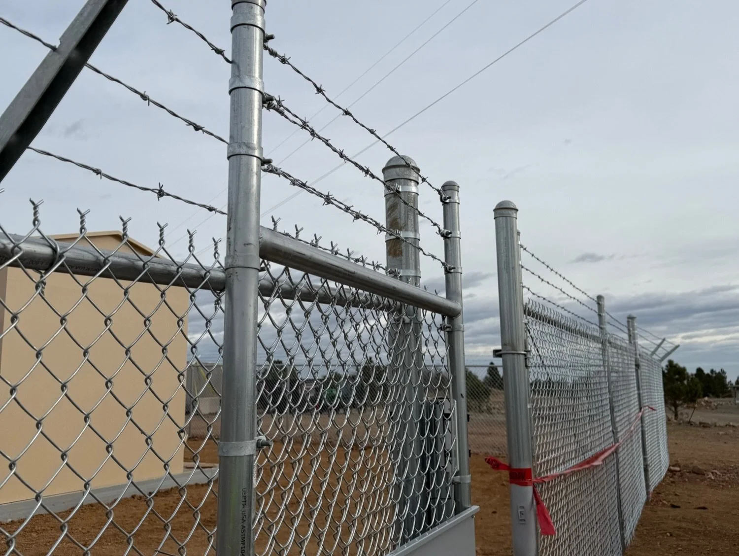 A chain-link fence with barbed wire on top, partially tied off with red tape, in a desert area with a beige building and cloudy sky in the background.