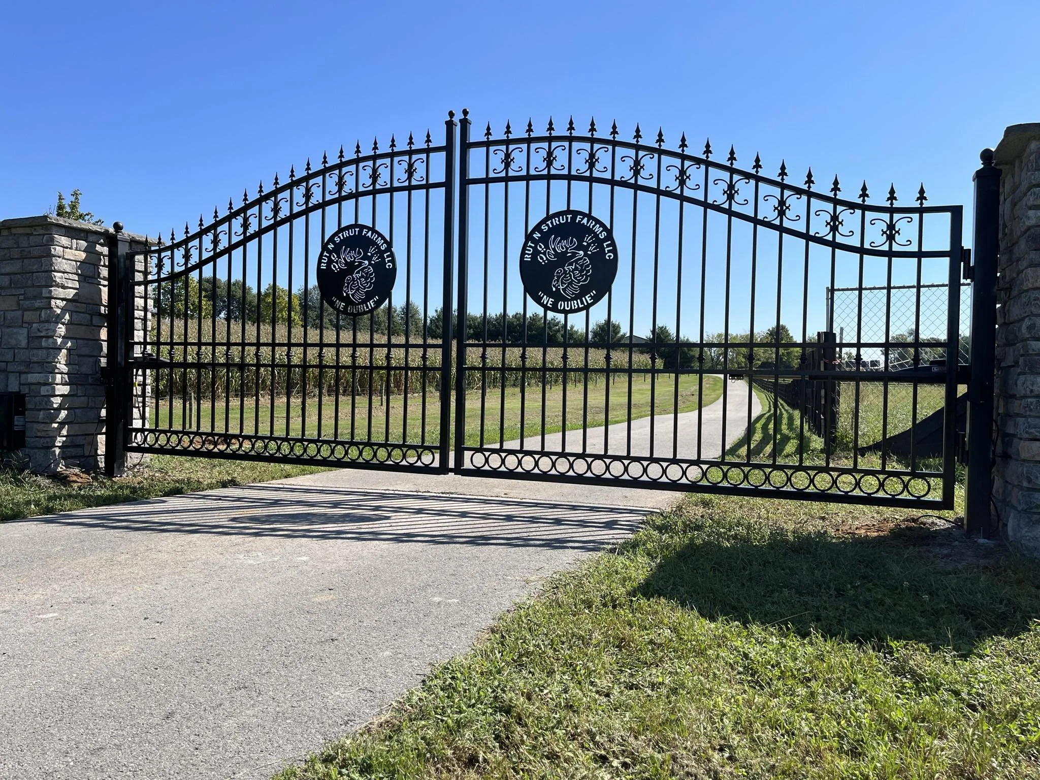 A black wrought iron gate with decorative accents and two round emblems featuring a devil and the text 'RUTN STRUT FARMS LLC' and 'INE OUBLIE.' The gate is set between stone pillars and is part of a farm entrance with a paved driveway and green field in the background.