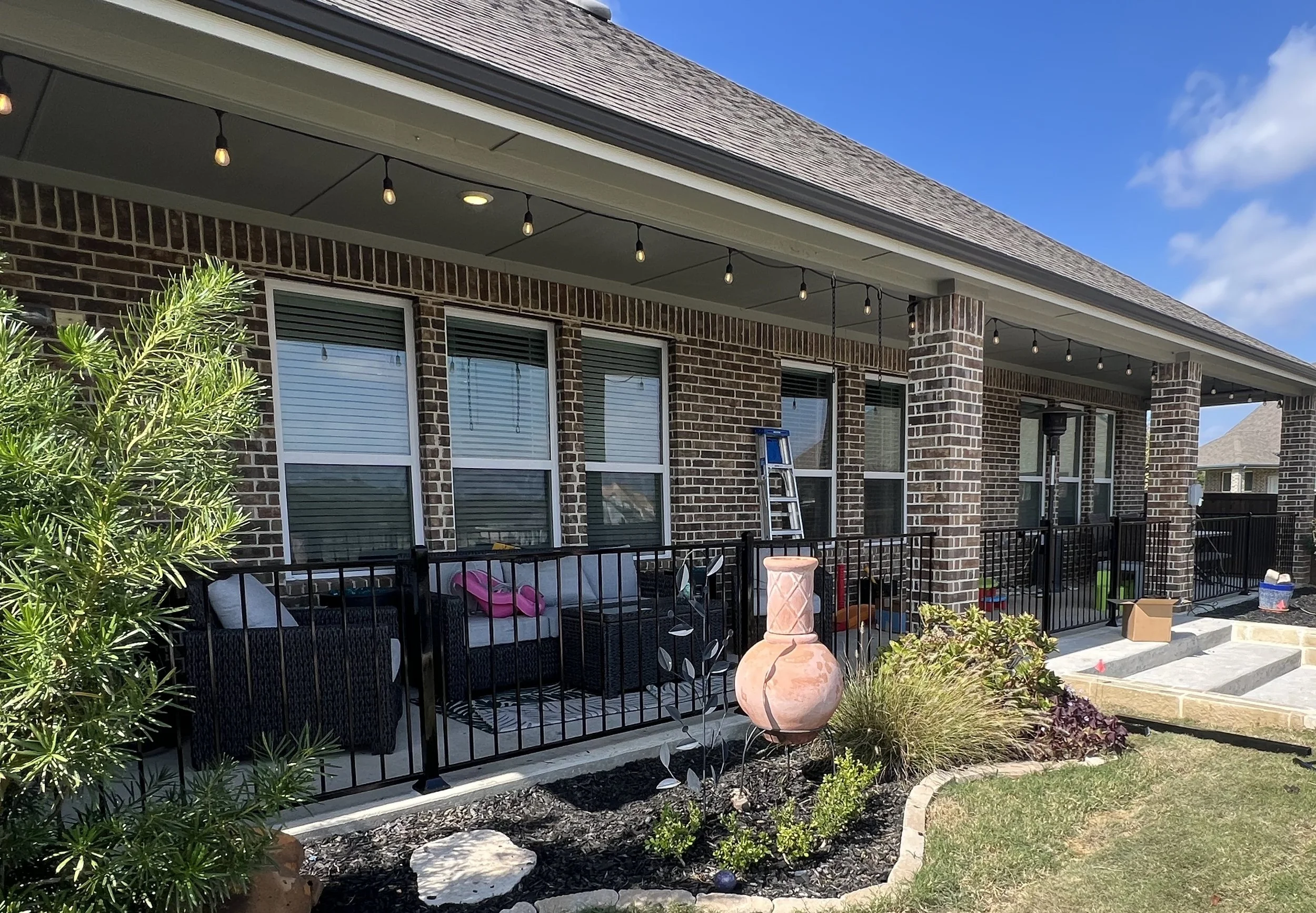 Back porch of a brick house with outdoor seating, string lights, and plants. Ladder leaning against the house. Steps leading to the porch with some boxes and objects nearby.