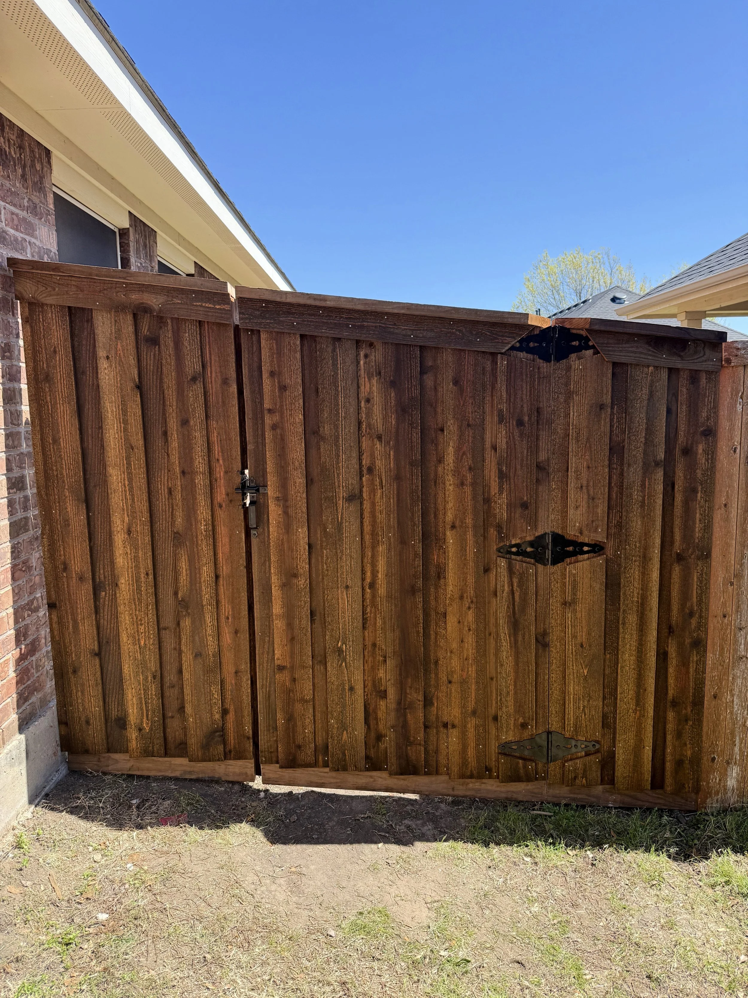 A wooden backyard gate with black hinges and latch, partially open, in front of a house with brick and beige siding, under a clear blue sky.