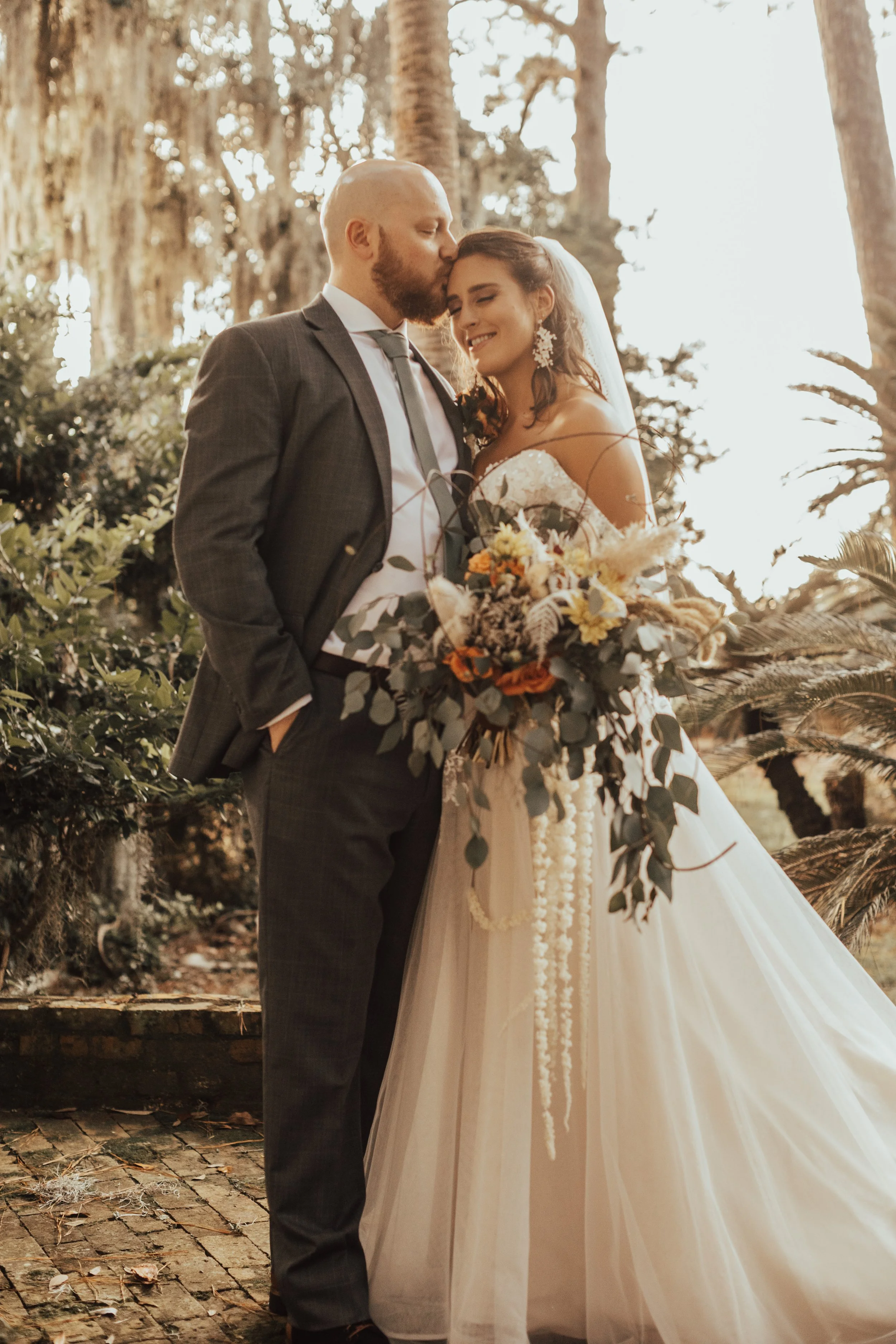 A bride and groom standing outdoors in a wooded area, with the groom kissing the bride on the forehead. The bride is holding a large bouquet of flowers and is smiling, dressed in a white strapless gown with a veil, while the groom is dressed in a gray suit with a tie.