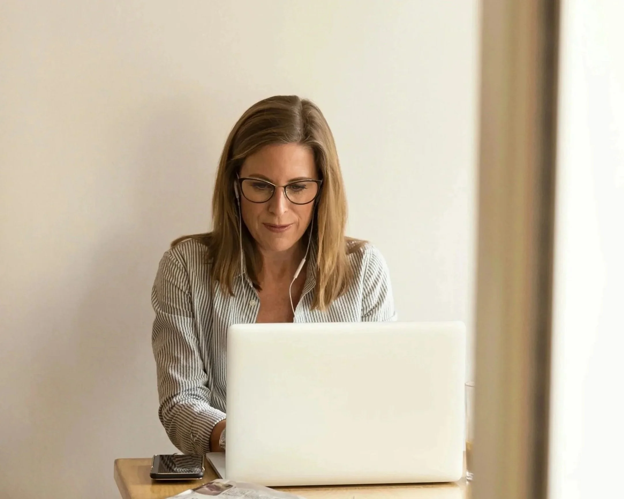 Woman with shoulder-length brown hair, glasses, wearing a striped shirt, sitting at a table and working on a laptop, with a smartphone nearby, in a well-lit room.