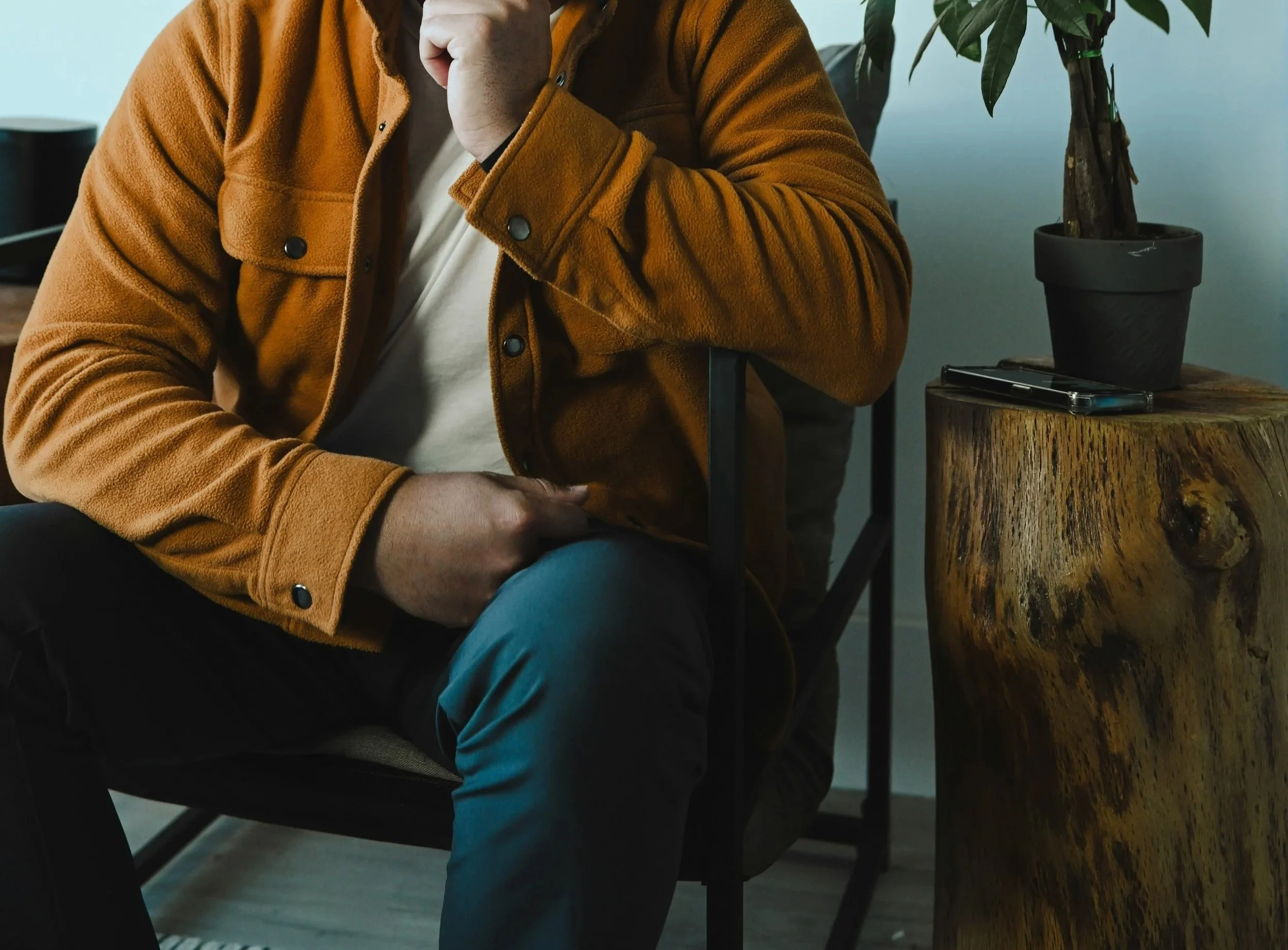 A person sitting on a chair, wearing a brown jacket over a white shirt, with their hand resting on their lap, and their other hand near their face. There is a wooden side table with a potted plant and a smartphone on it.