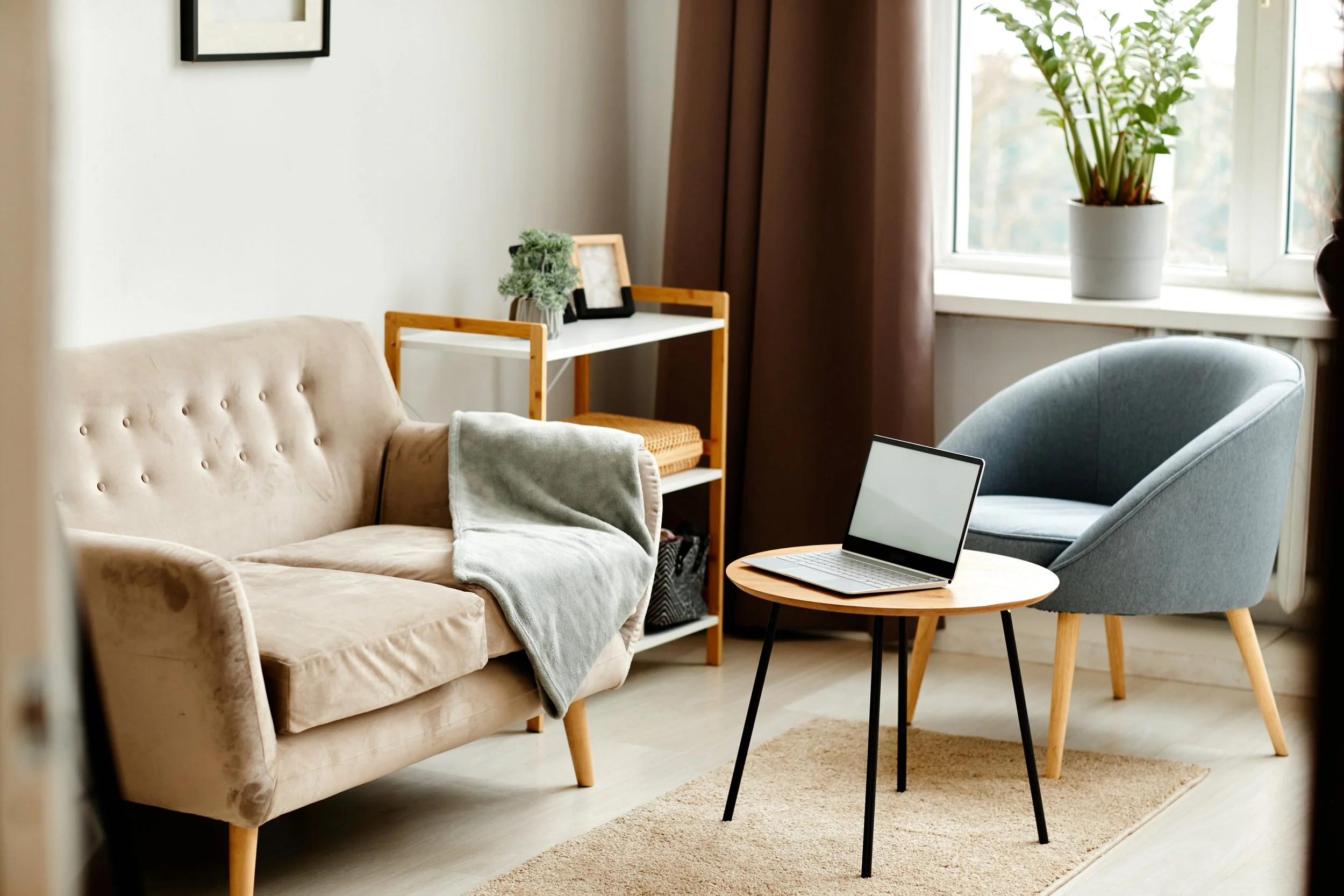 Living room with beige sofa, gray armchair, small wooden table with a laptop, white shelf with plants and picture frames, and a window with potted plants and brown curtains.