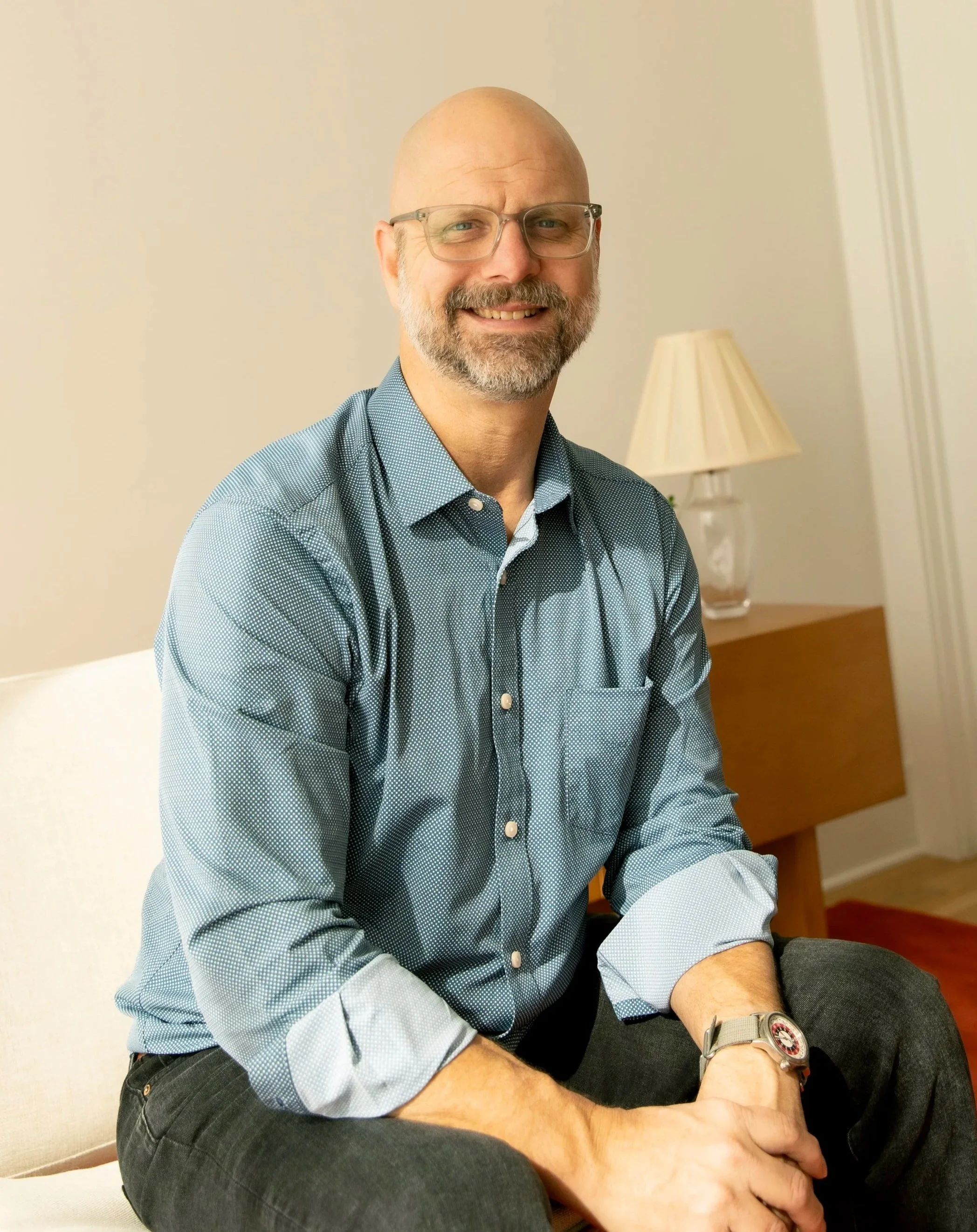 A middle-aged man with glasses, a beard, and a shaved head sitting on a couch in a living room. He is smiling and wearing a blue patterned button-up shirt with rolled-up sleeves, dark pants, and a wristwatch. There is a table with a lamp behind him.