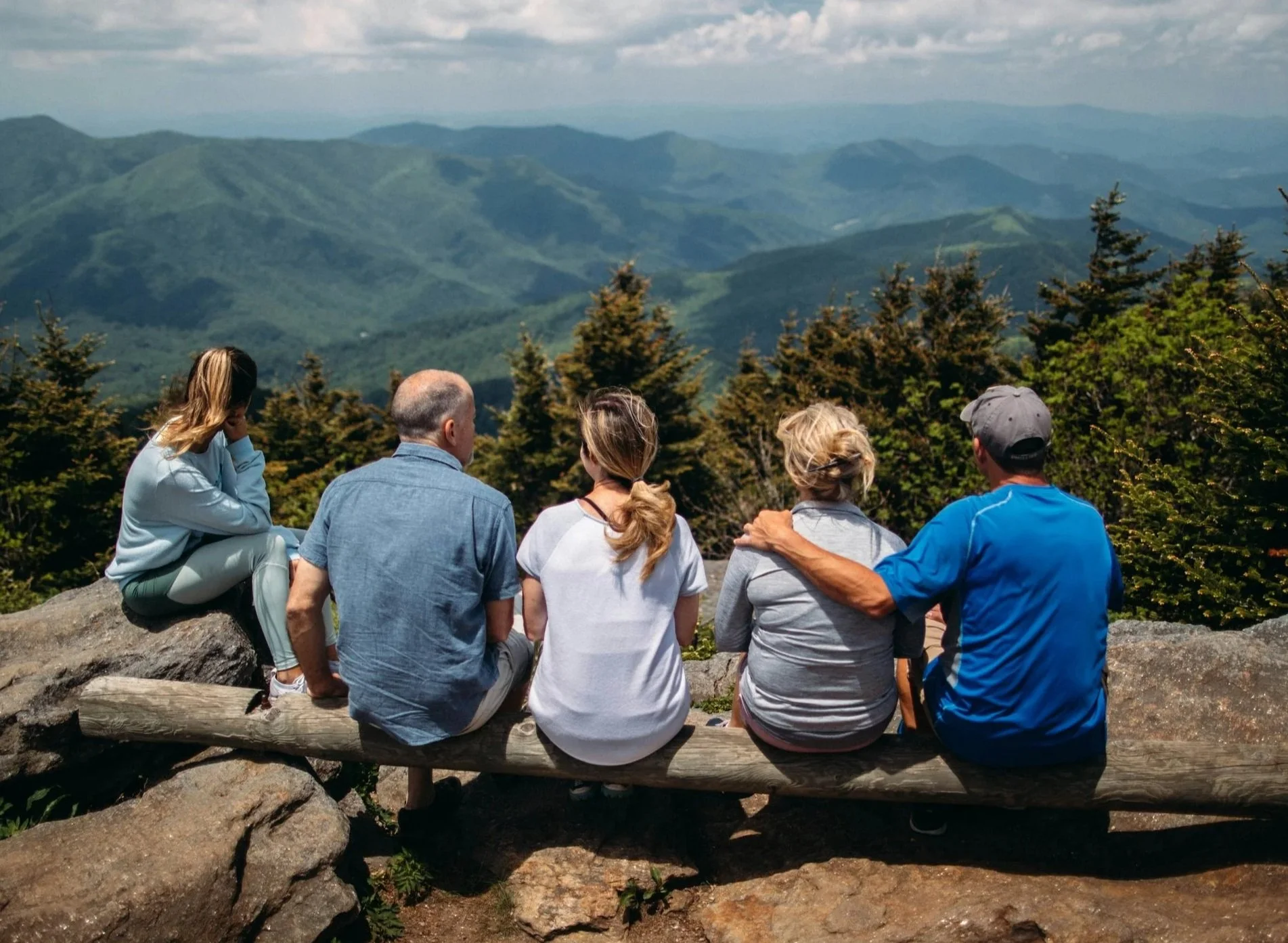 A group of five people sitting on a log on a mountain, overlooking a lush green mountain landscape.