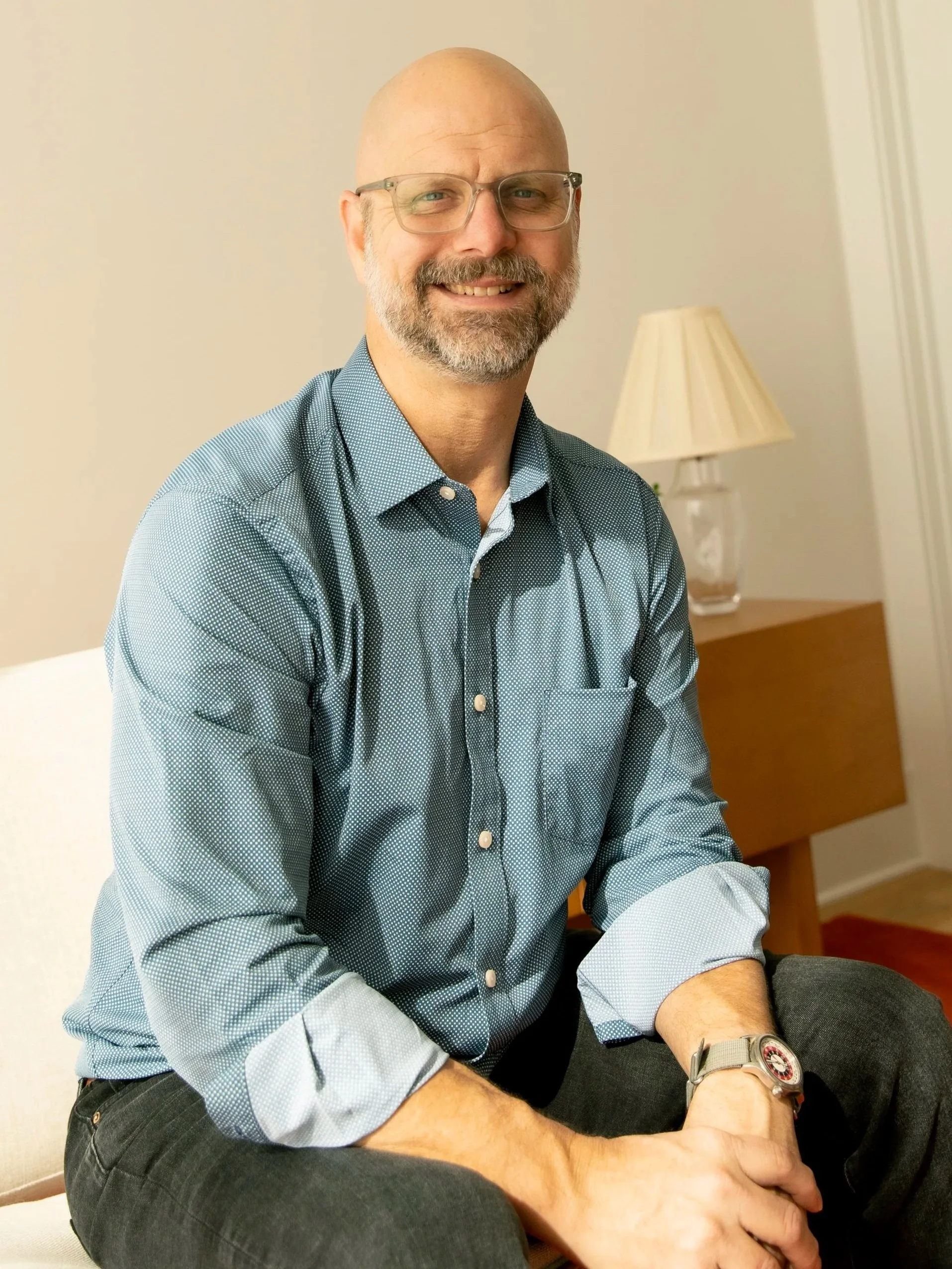 A smiling middle-aged man with glasses, a beard, and a bald head, wearing a blue patterned button-up shirt with rolled-up sleeves, sitting on a white chair in a room with a wooden side table and a lamp in the background.