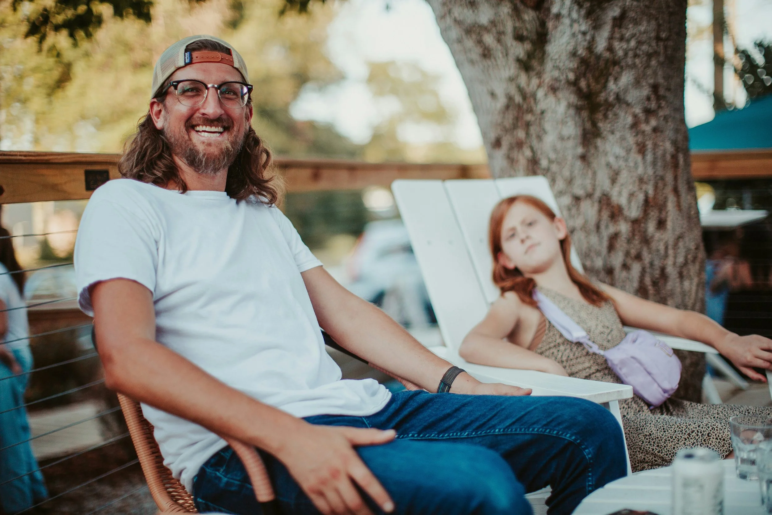 A man with long hair, glasses, and a beard, smiling while sitting outdoors in a white t-shirt and a trucker hat, with a young girl relaxing on a lounge chair next to him under a large tree.